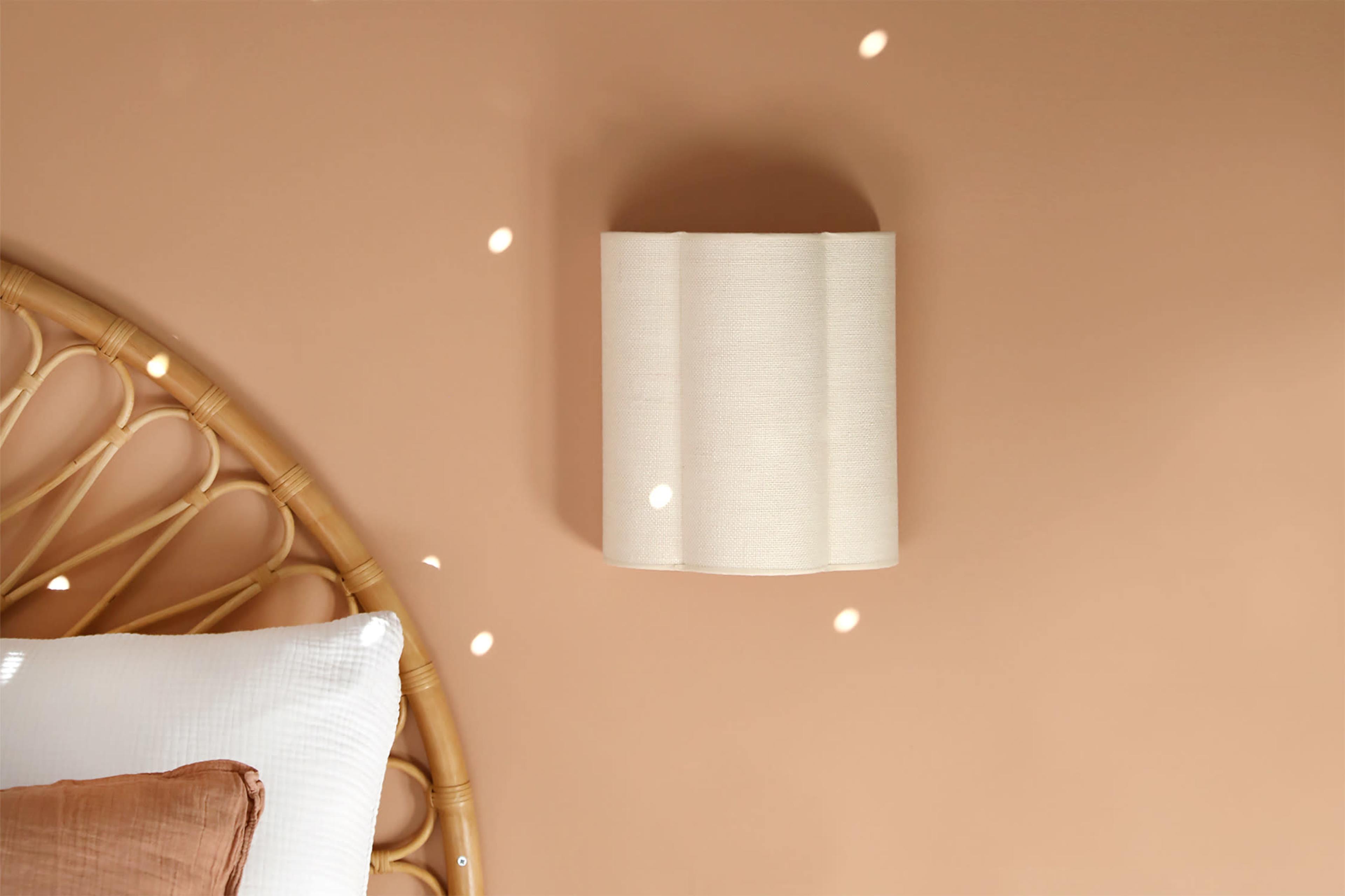 A soft, beige wall lamp is mounted on a light brown wall beside a rattan bed headboard with white bedding.