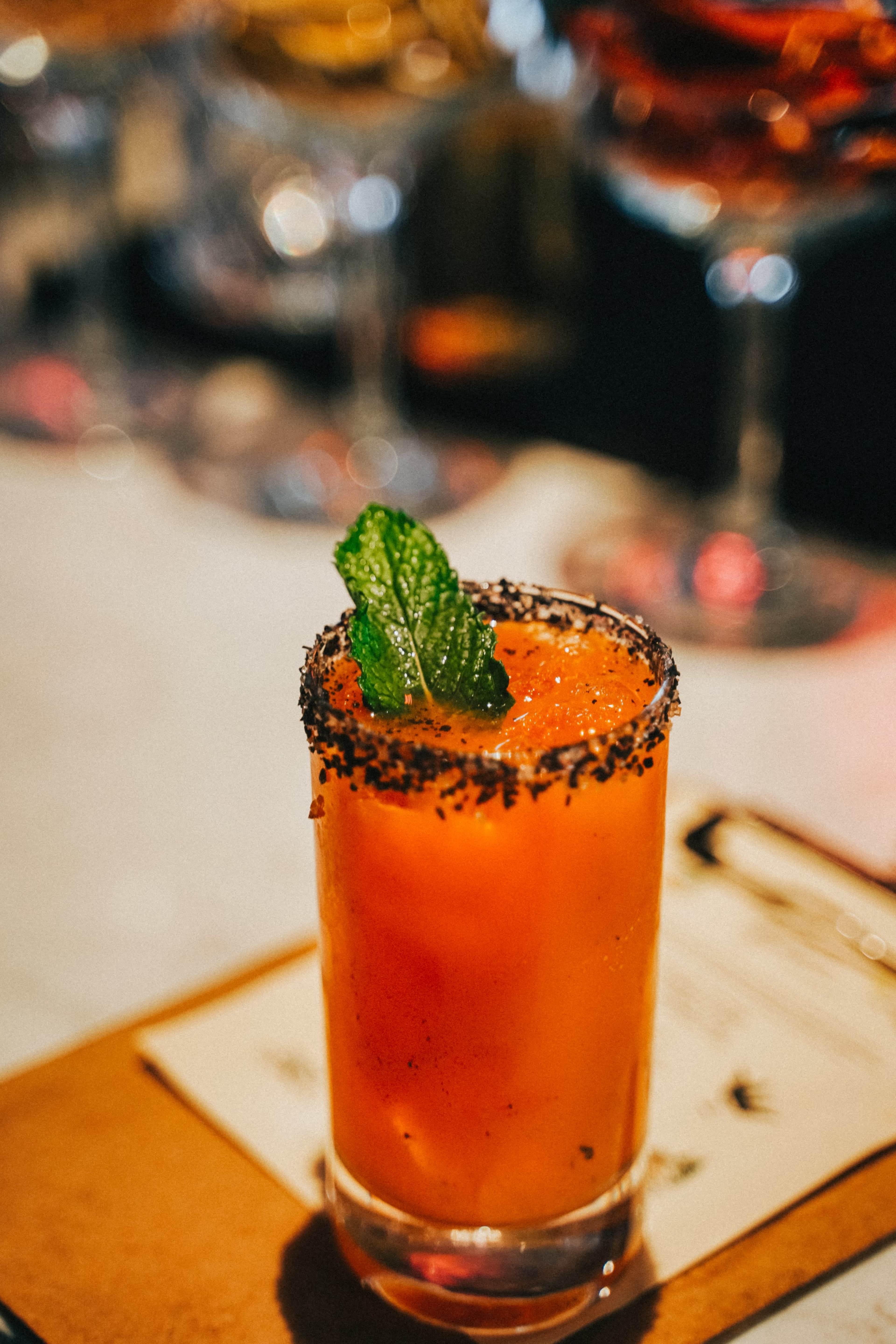 A glass of vibrant orange juice garnished with a mint leaf and a black salt rim sits on a wooden surface.