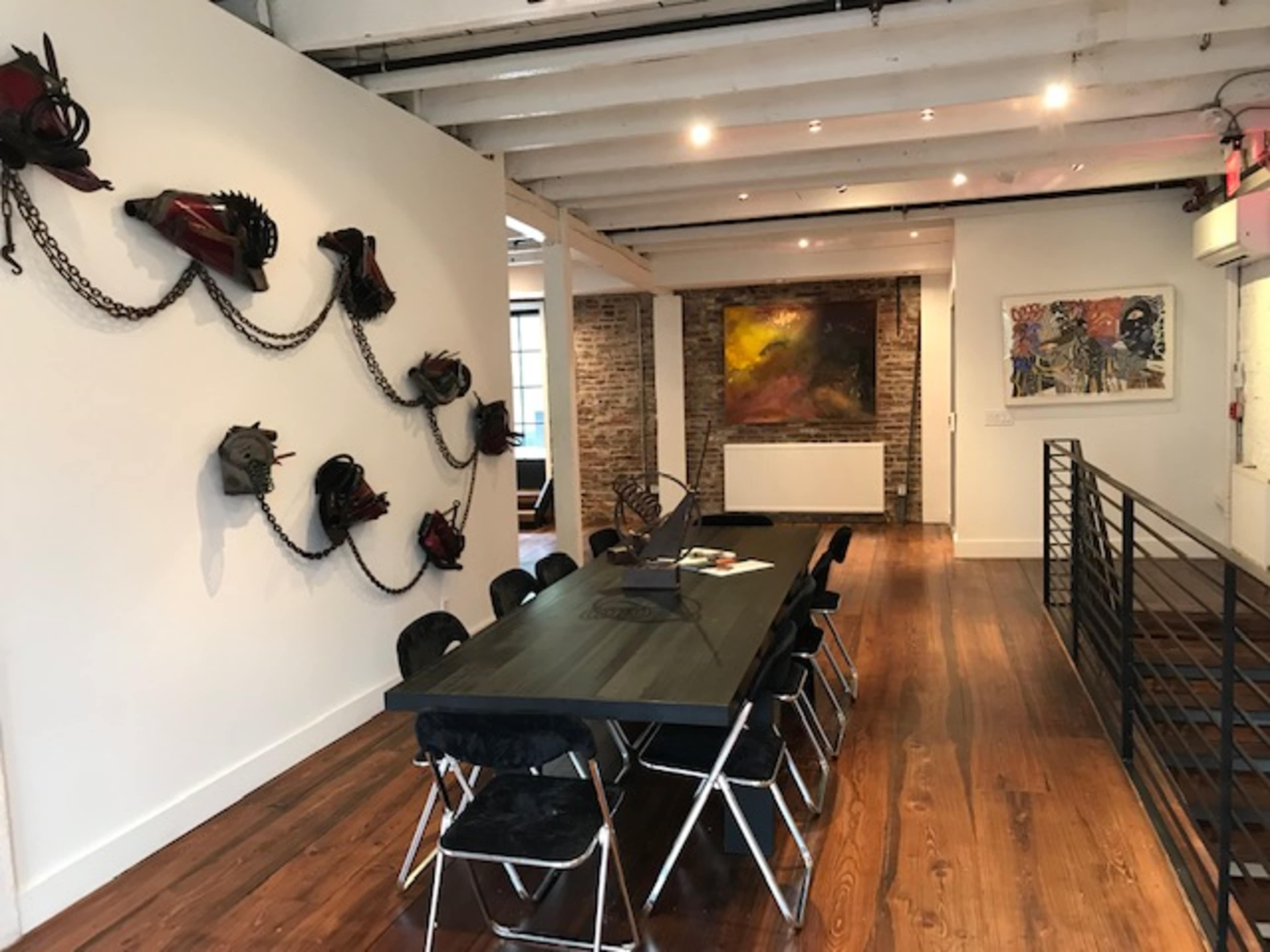 The image shows a modern interior space featuring a long black table surrounded by folding chairs, with artistic wall decor and a painting in the background.
