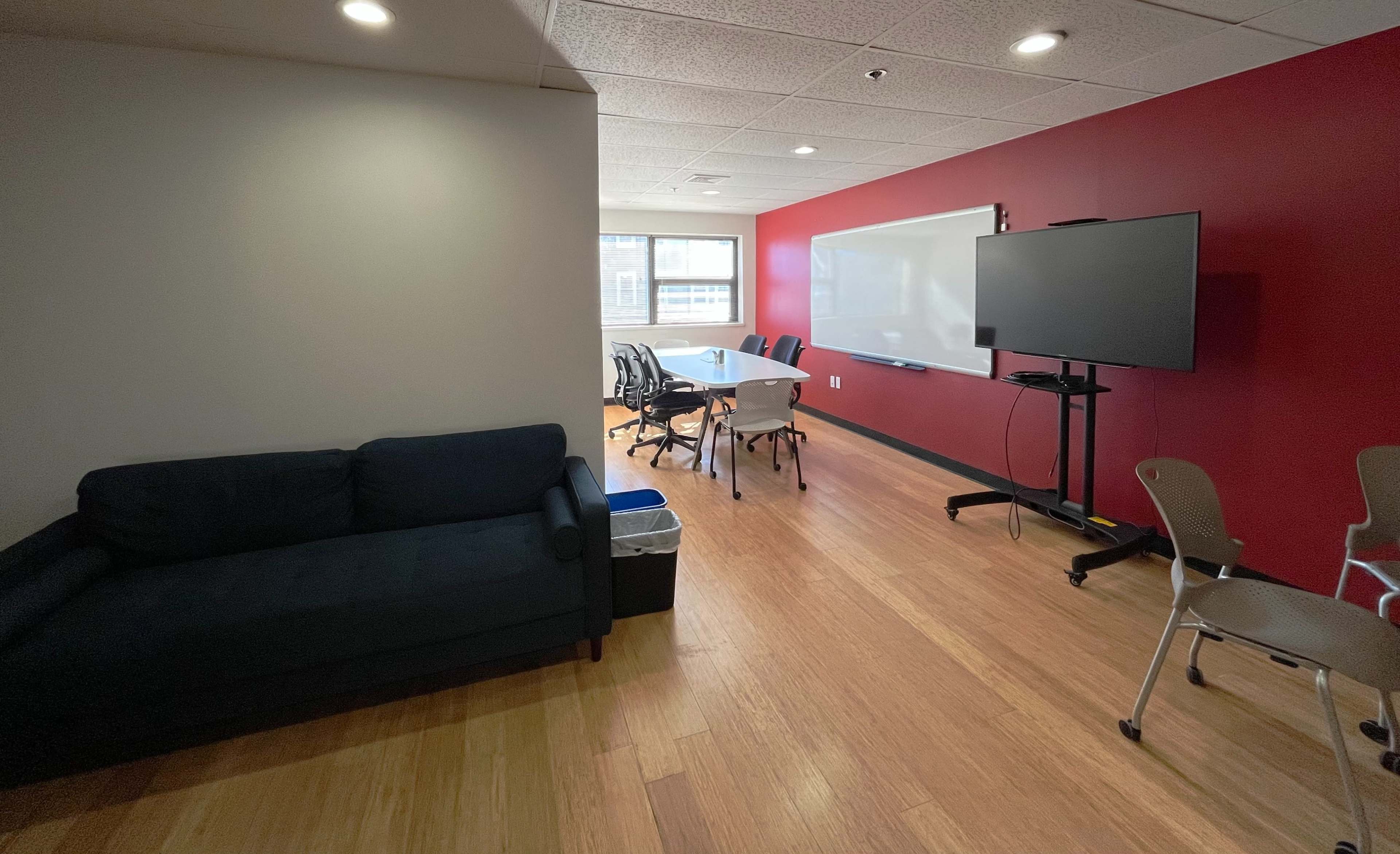 The image shows a meeting room with a table and chairs, a large screen on a stand, and a dark couch near a wall.