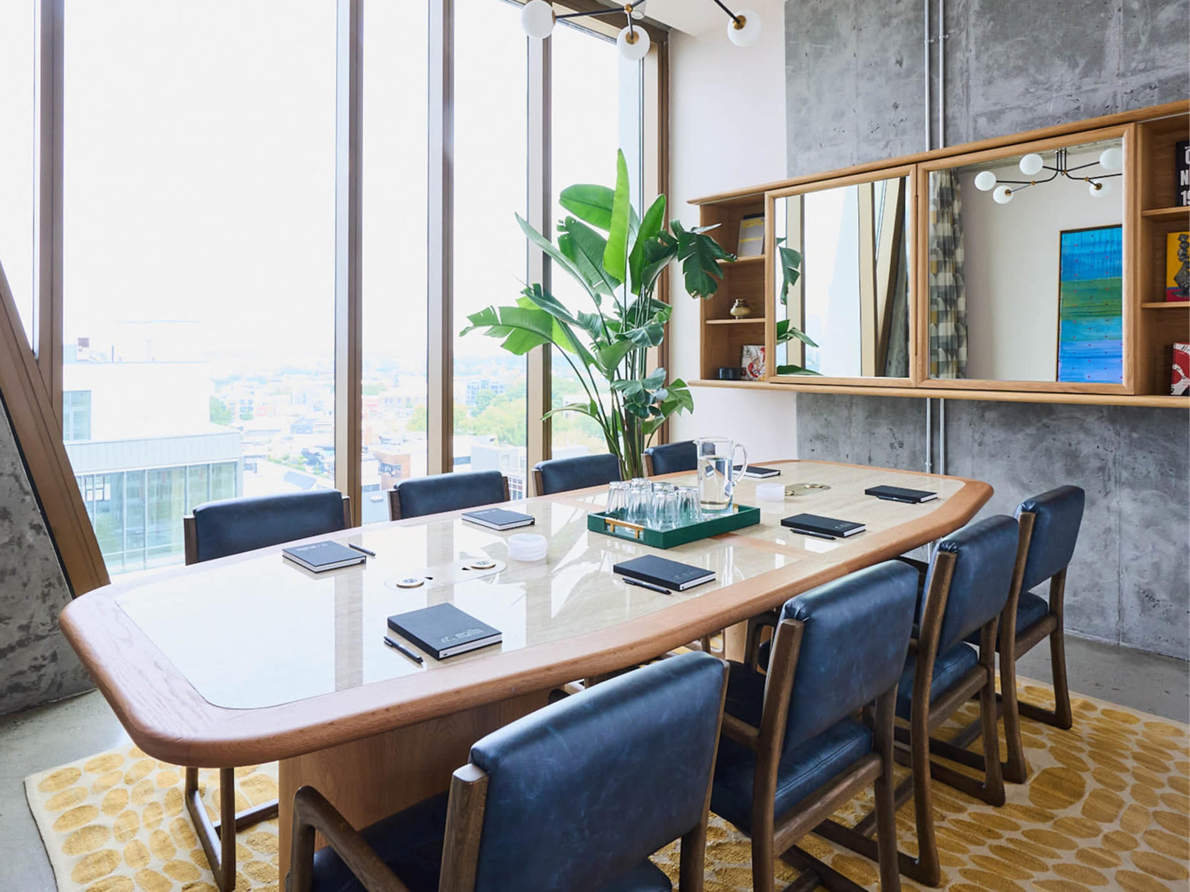 The image shows a modern conference room with a large wooden table surrounded by blue chairs, large windows offering a city view, and a potted plant in the corner.