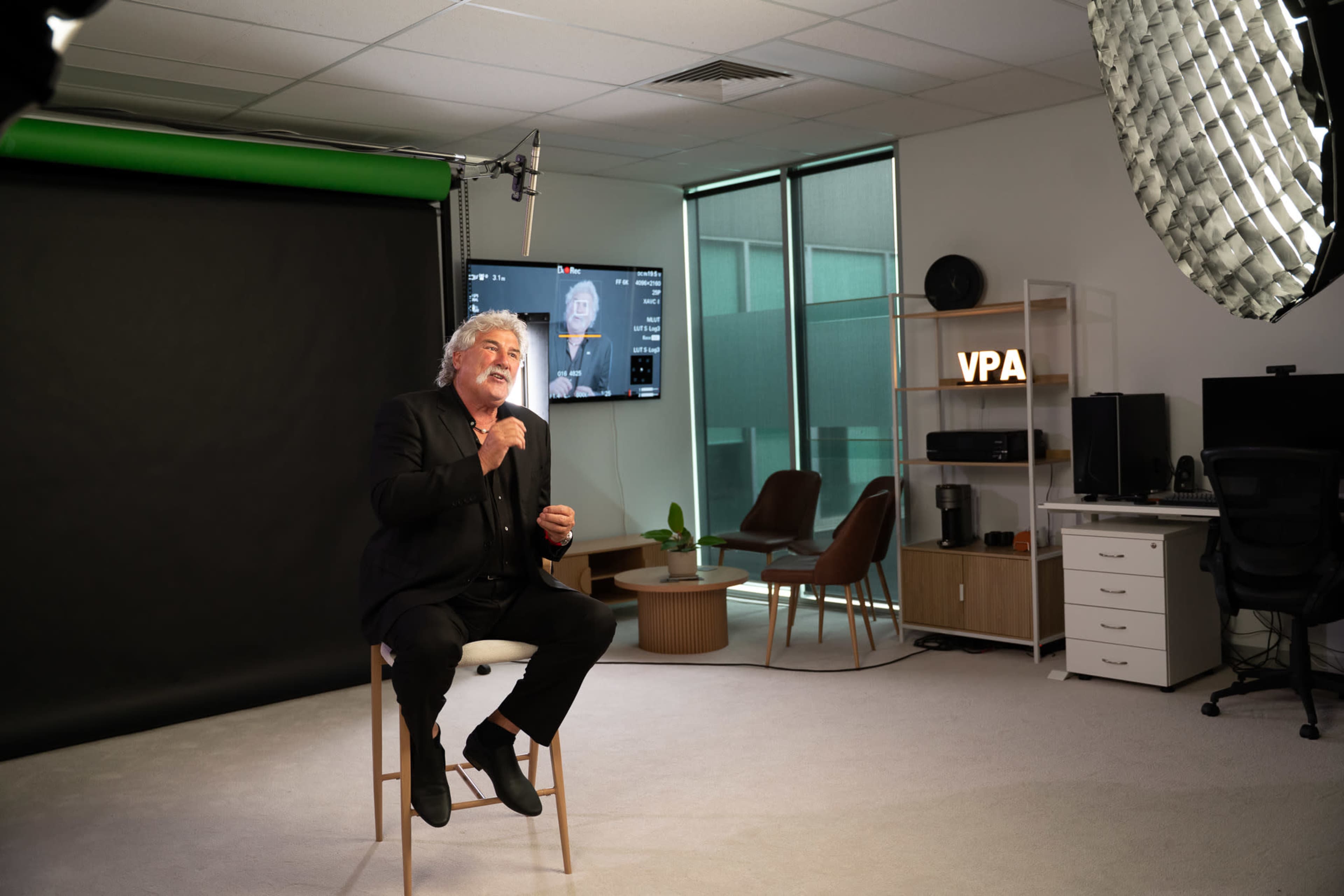 A man is seated on a stool in a well-lit studio, speaking while a camera setup captures the scene in front of a black backdrop.