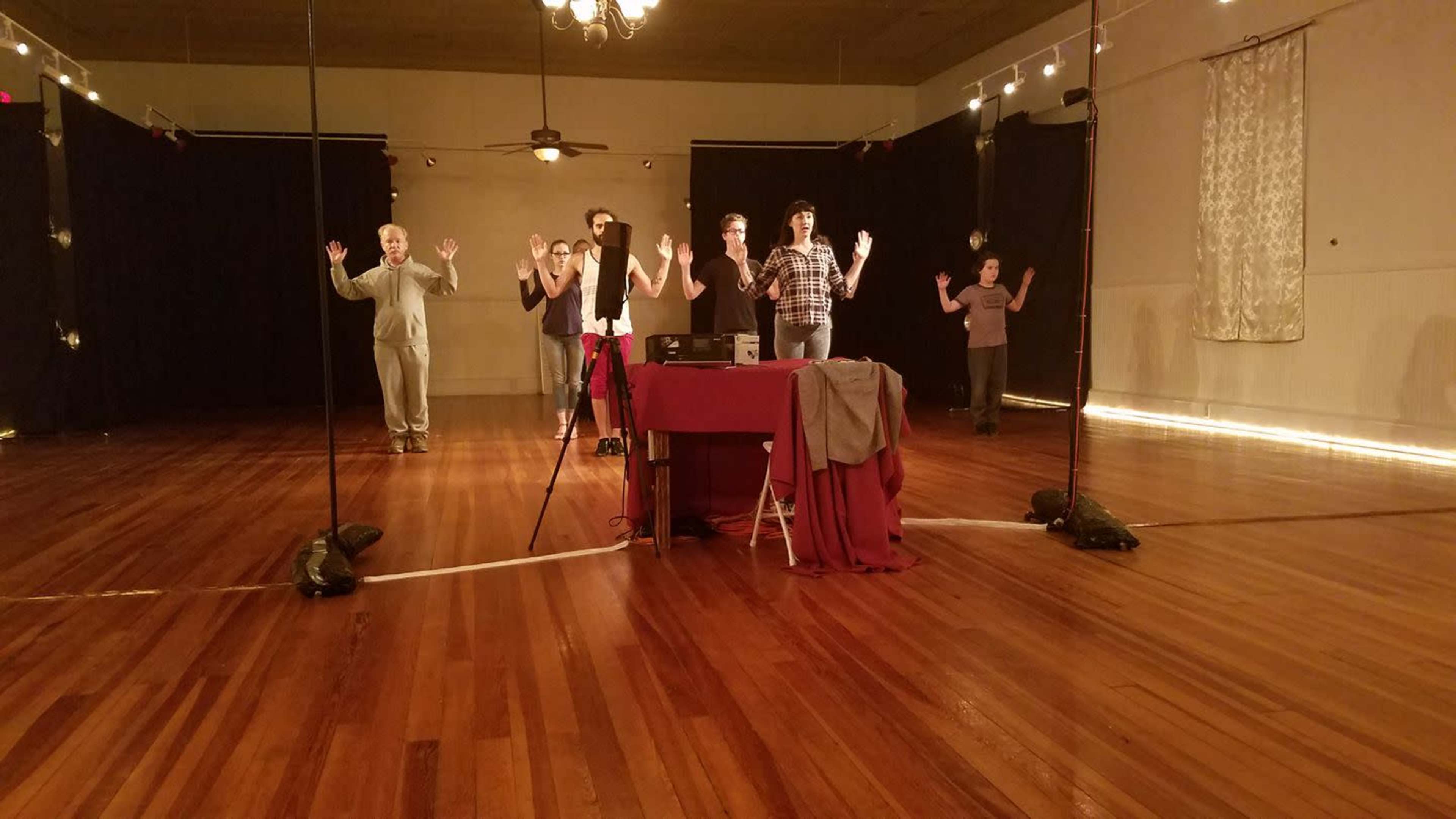 A group of six people stands with their hands raised in a room with wooden floors and wall mirrors, while a table covered in a red cloth is positioned in the foreground.