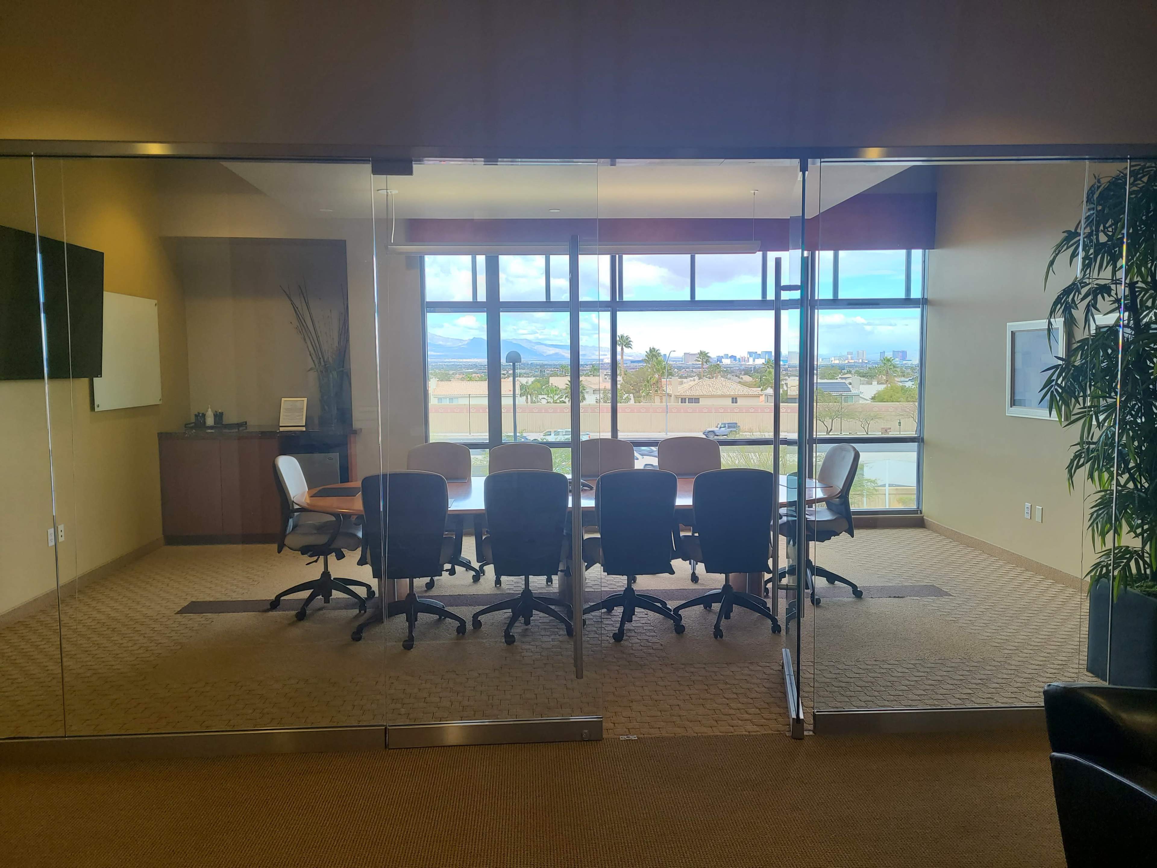 A spacious conference room with a large table and rolling chairs, featuring a glass wall that offers a view of the cityscape and mountains beyond.
