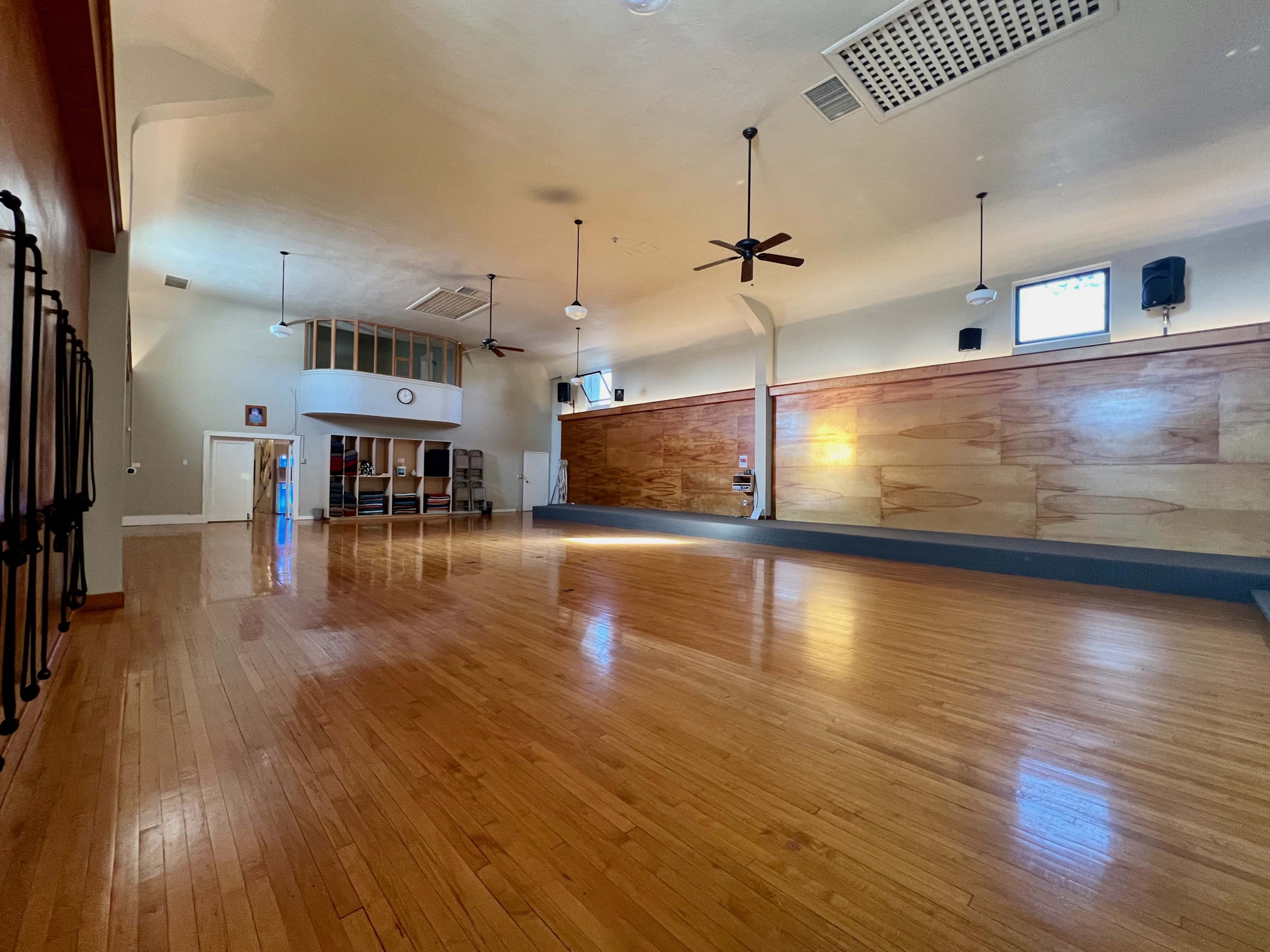 A spacious, well-lit wooden gym or studio with a polished floor and tall ceilings, featuring fans and large wooden wall panels.