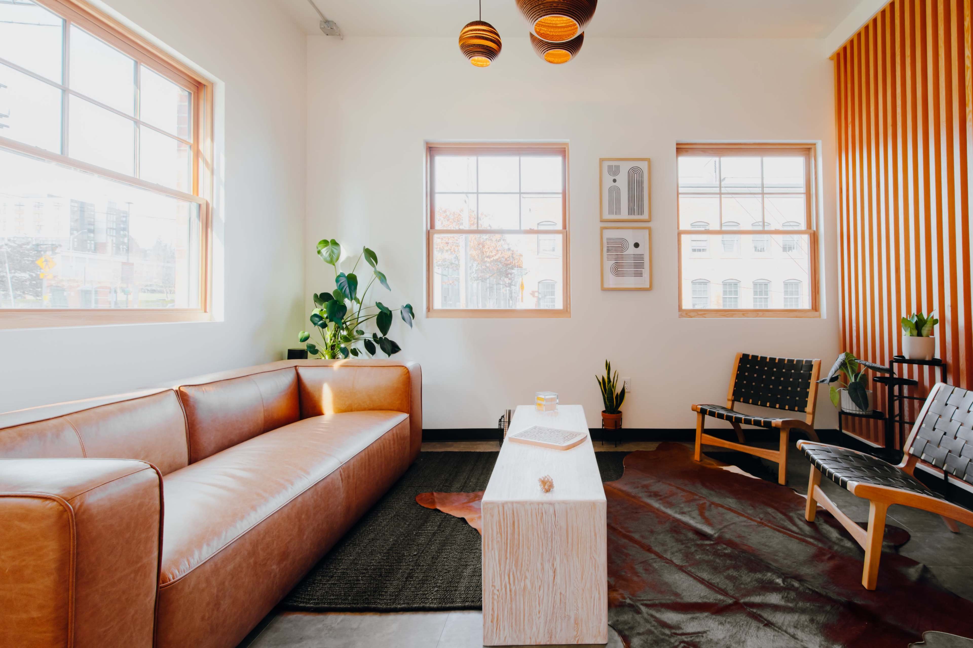A modern living room featuring a brown leather sofa, a wooden coffee table, and various plants near large windows.