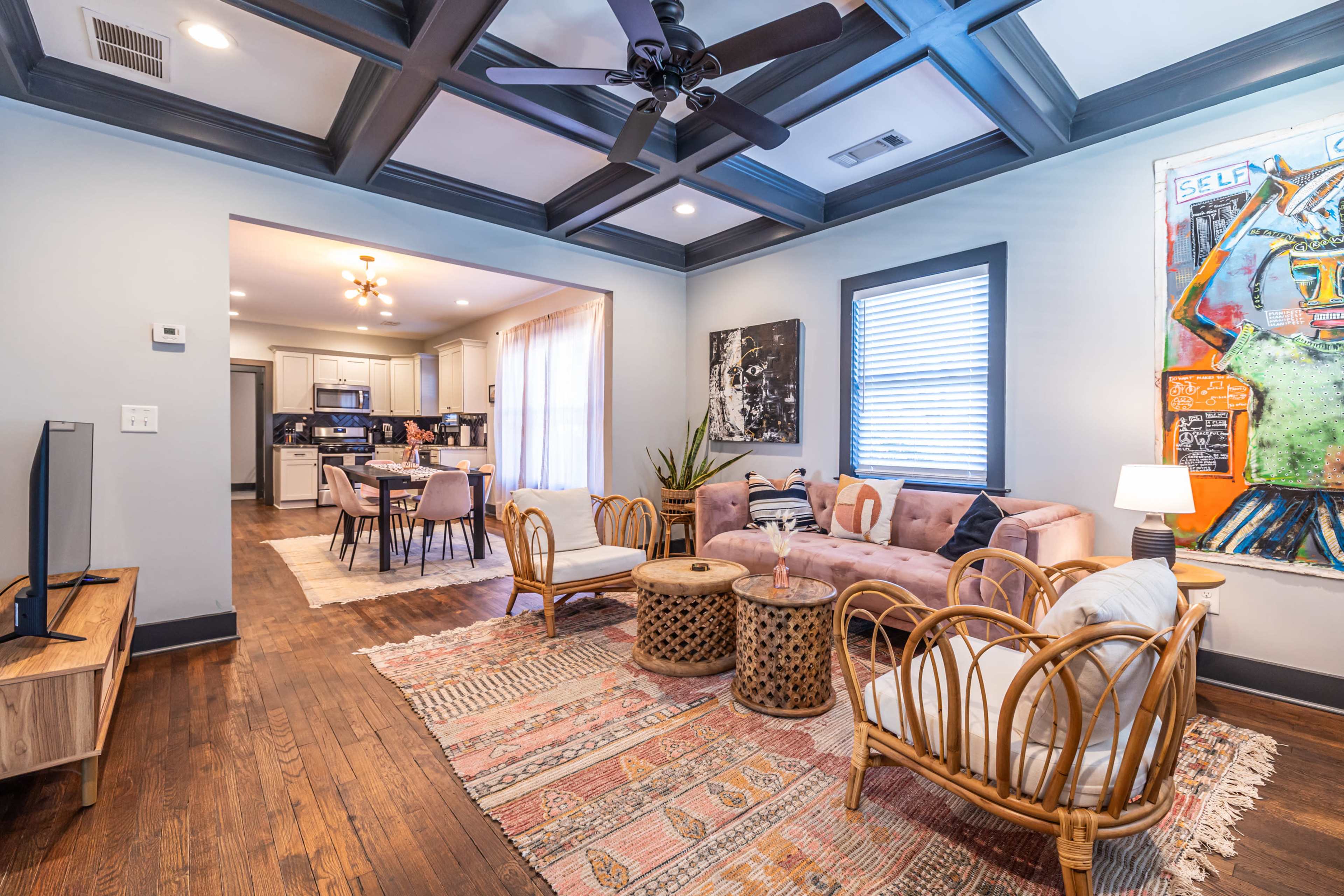 The living room features a pink sofa and rattan chairs, with a large painting on the wall and a view into a kitchen area.