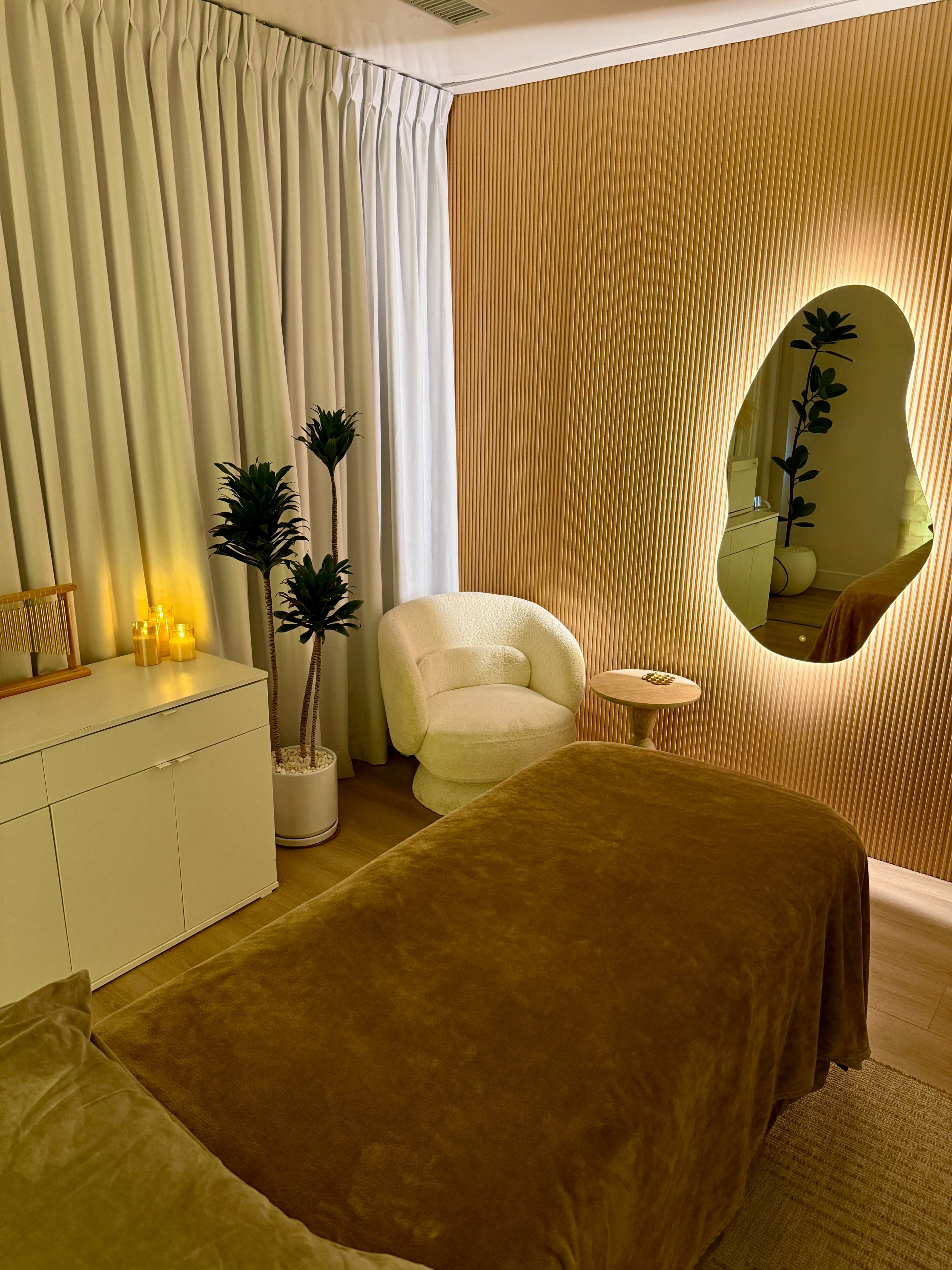 The scene features a minimalist room with a bed covered in a brown textile, a cozy chair, a small side table, and a backlit mirror against a textured golden wall.