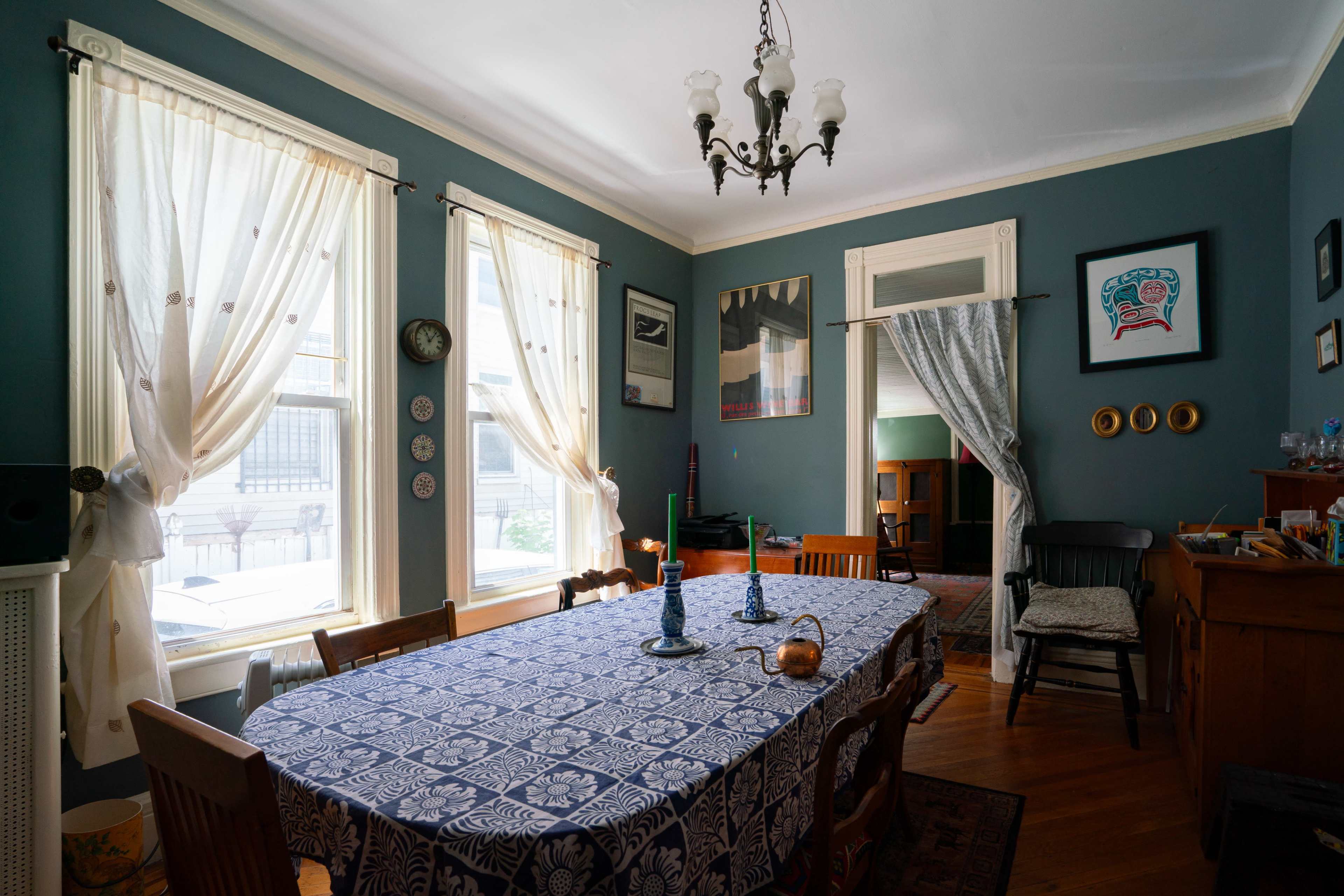 The image shows a dining room with a patterned tablecloth, a wooden table surrounded by chairs, and large windows adorned with sheer curtains.