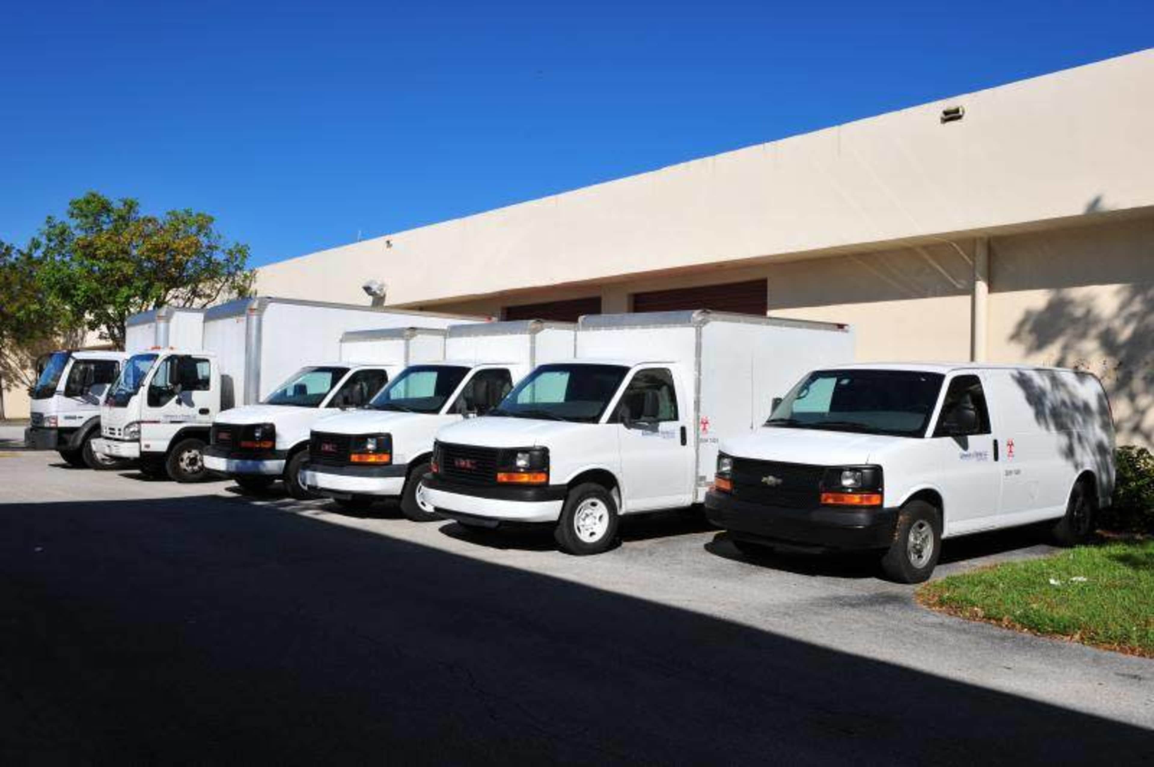 A line of six white delivery vans is parked beside a light-colored building.
