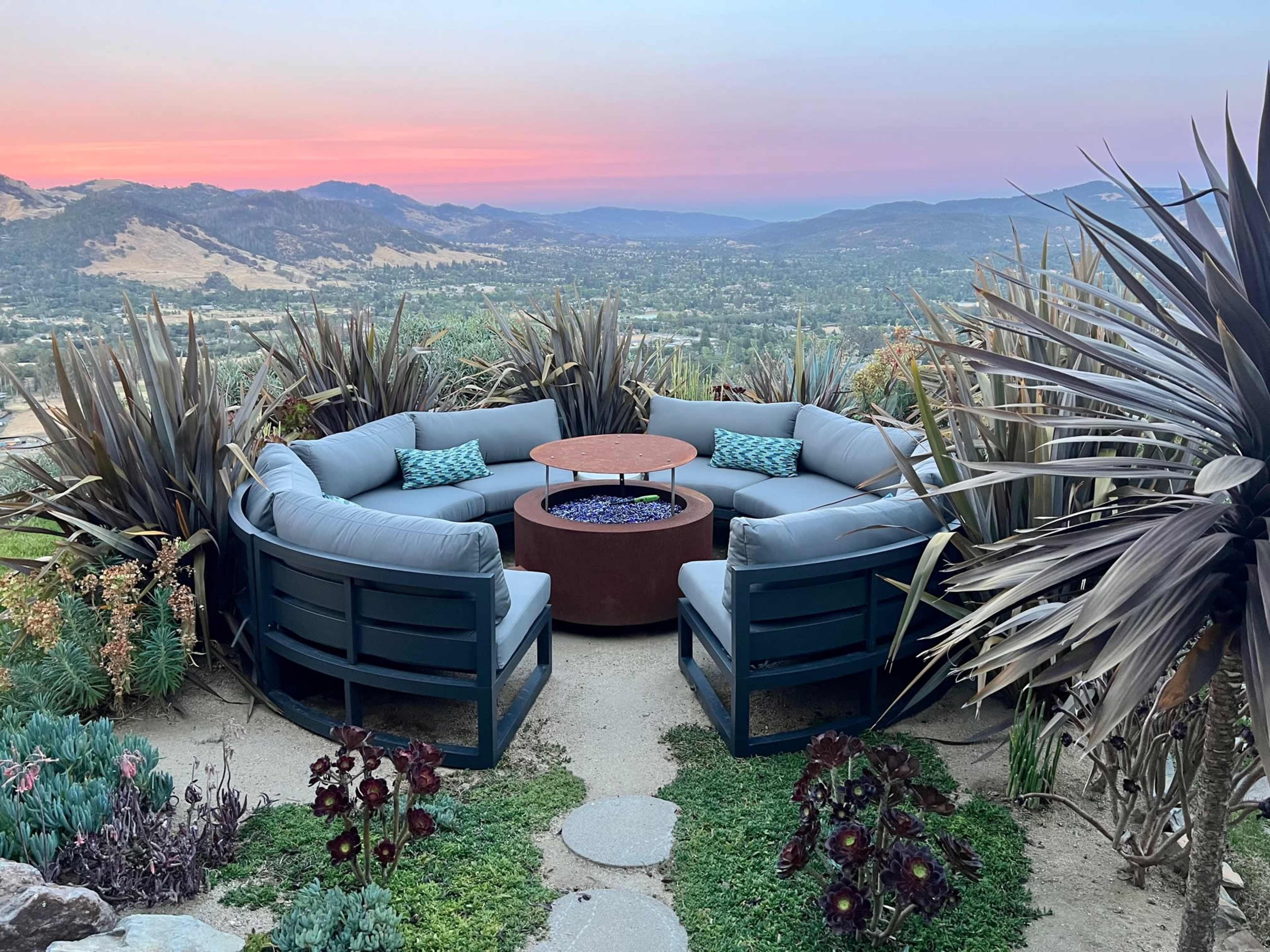 A circular seating area with blue cushions surrounds a central fire pit, set against a backdrop of mountains and valleys at sunset.