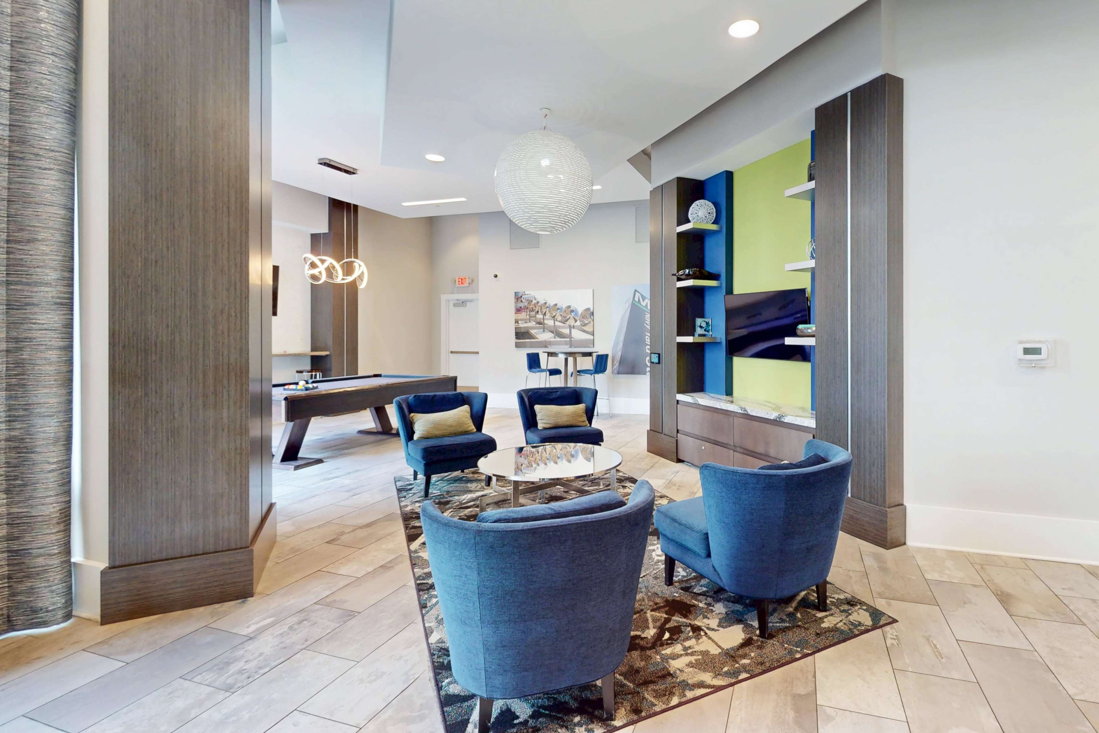 The image shows a modern lounge area with four blue chairs arranged around a circular table, featuring a decorative rug and a wall-mounted shelf with decorative items.