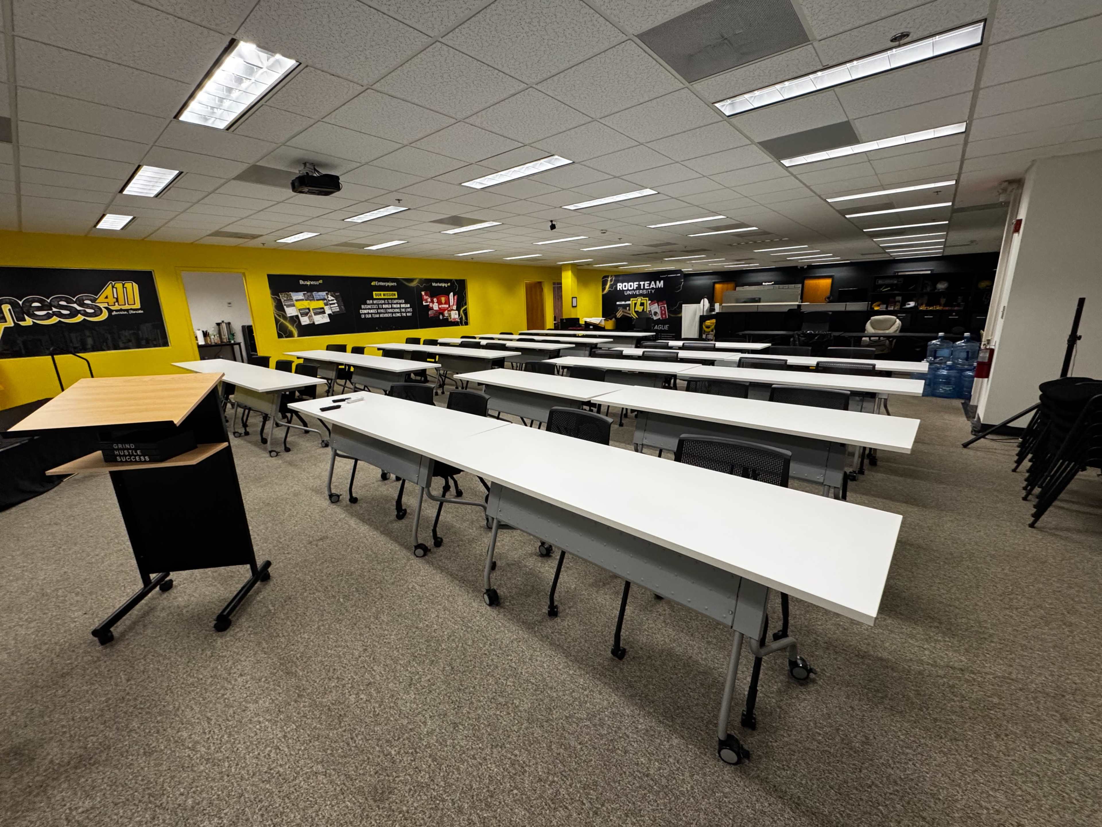 The image shows a spacious classroom or training room with multiple long, white tables arranged in rows, set against yellow and black walls decorated with printed materials.