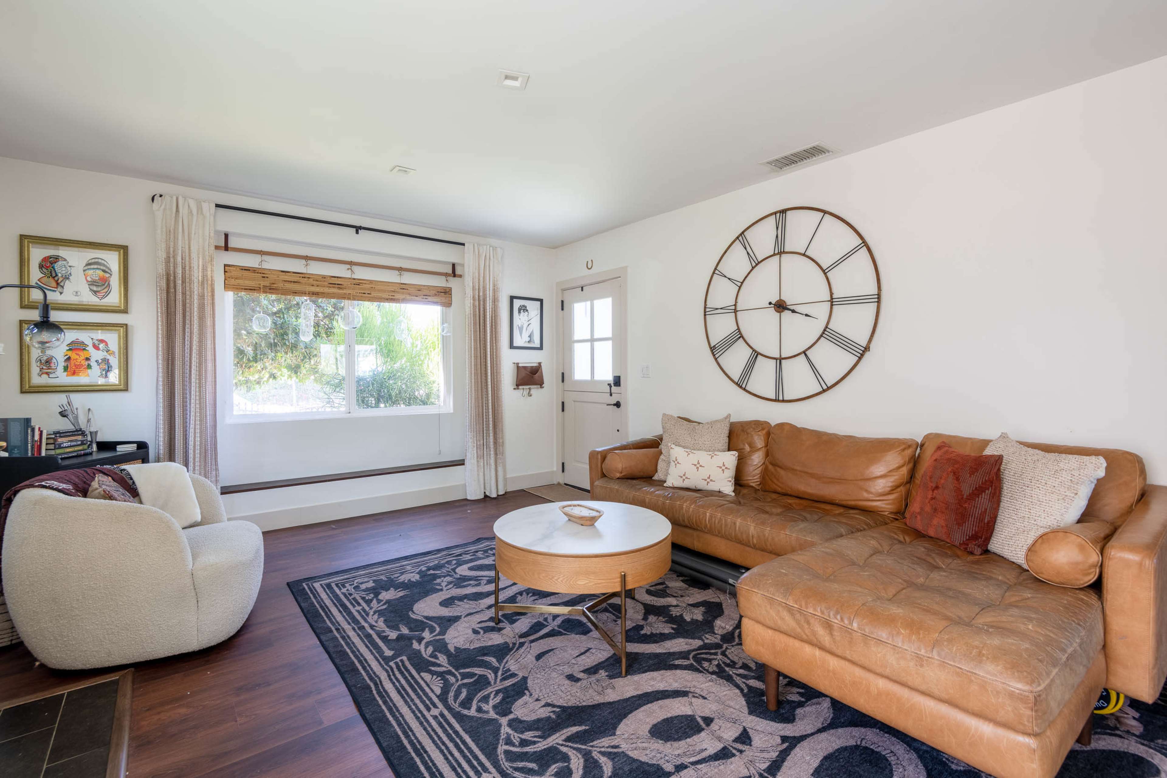 The image depicts a modern living room featuring a leather sectional sofa, a round coffee table, a large wall clock, and a window with wooden blinds.