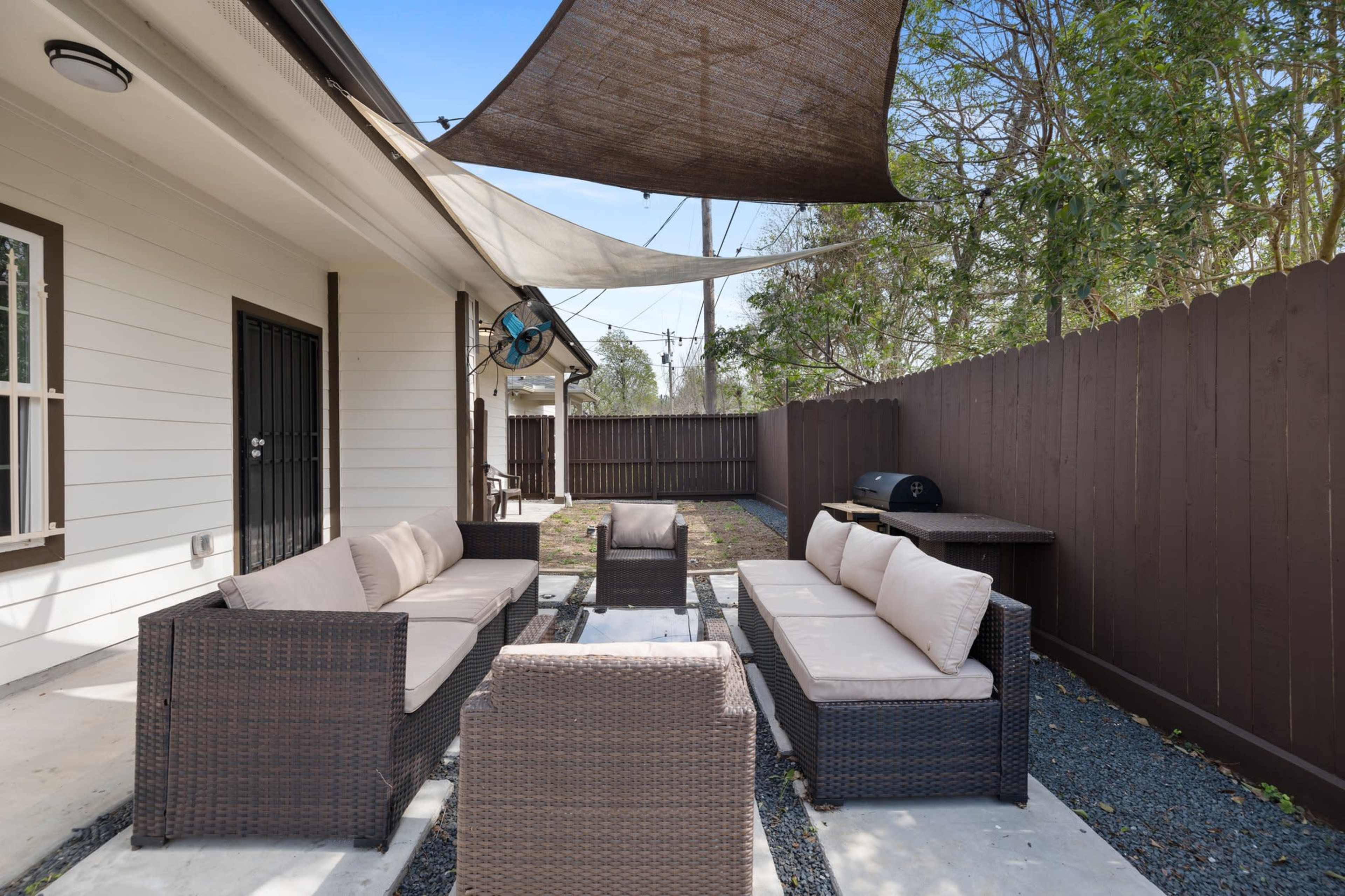 An outdoor seating area with a dark wicker sofa set, a glass-top table, a barbecue grill, and shaded by fabric canopies.