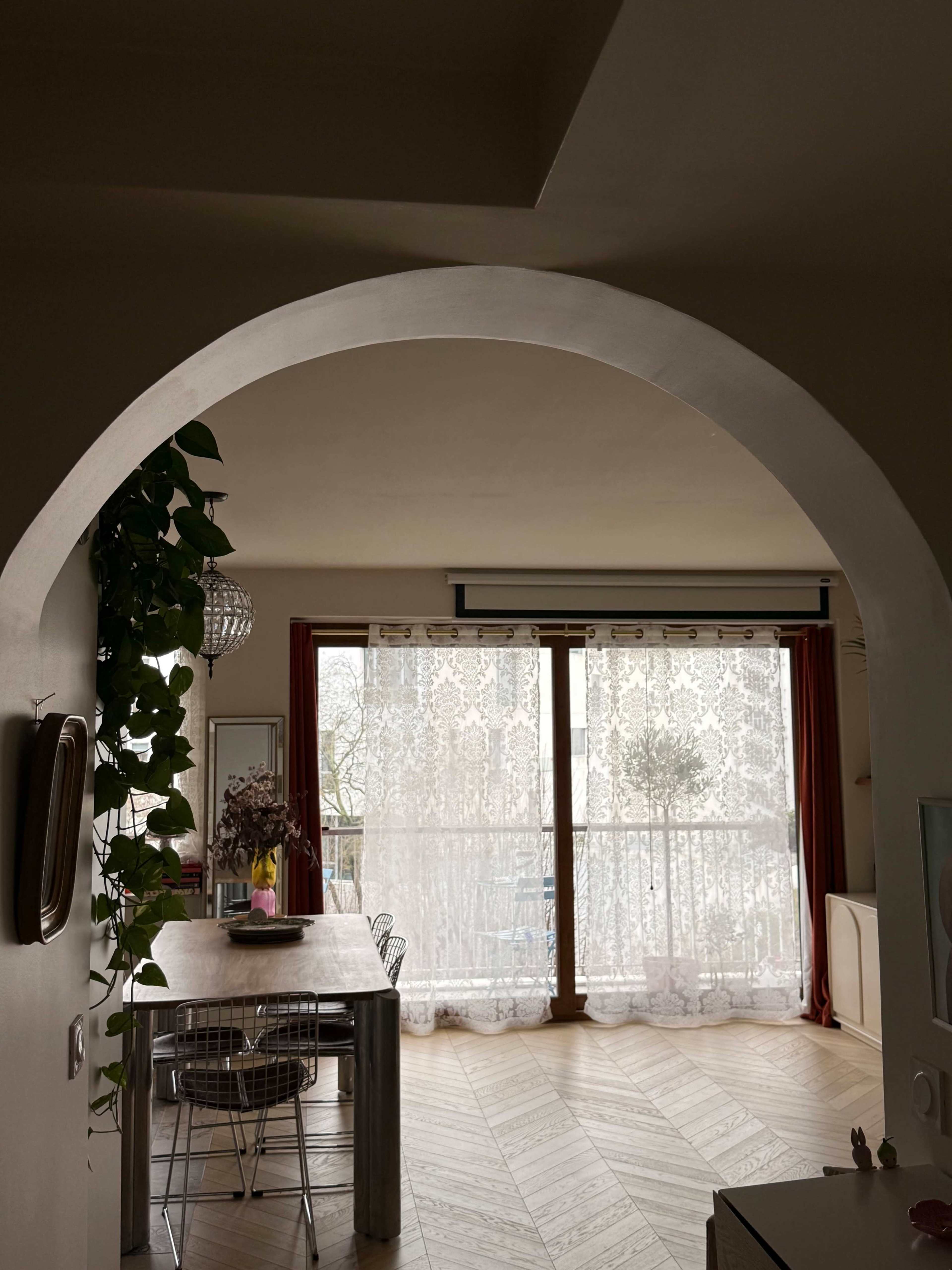 The image shows a dining area framed by an arch, featuring a wooden table, chairs, and large windows draped with sheer curtains that overlook a balcony.