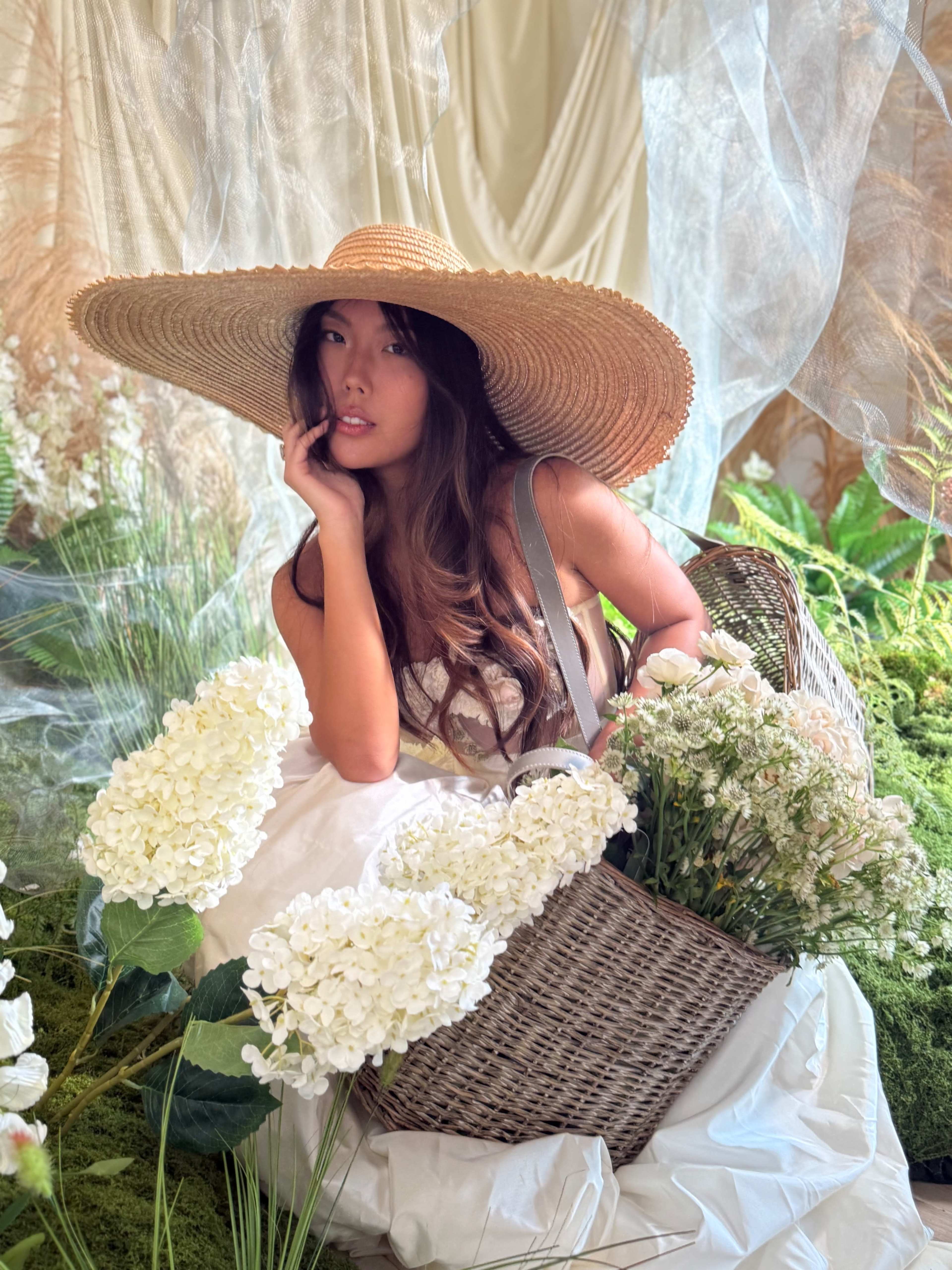 A woman wearing a large straw hat poses with a basket of flowers in a styled setting featuring draped fabrics and greenery.