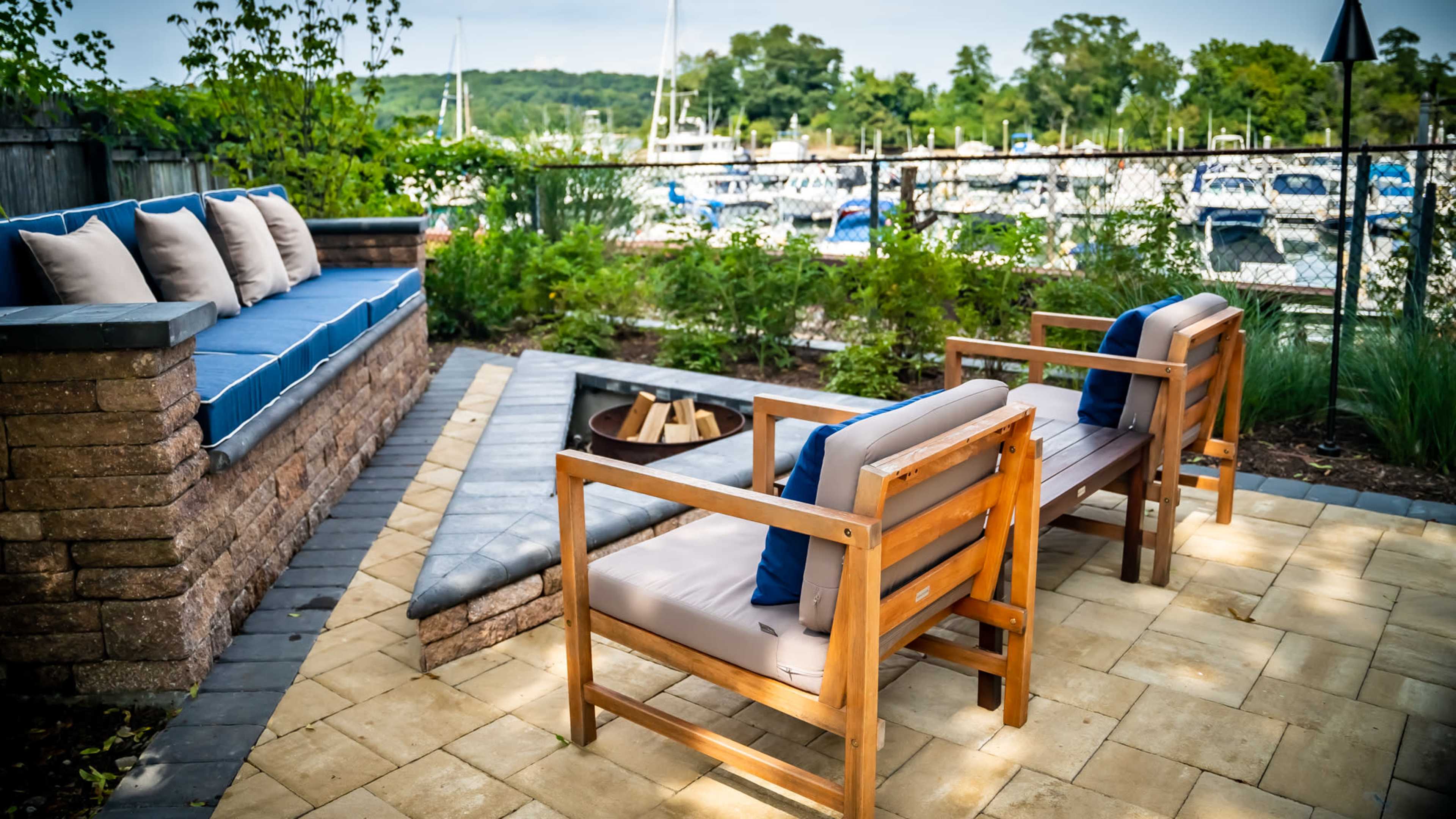 The image shows a patio area with two wooden chairs facing a fire pit and a stone seating wall, overlooking a marina filled with boats.