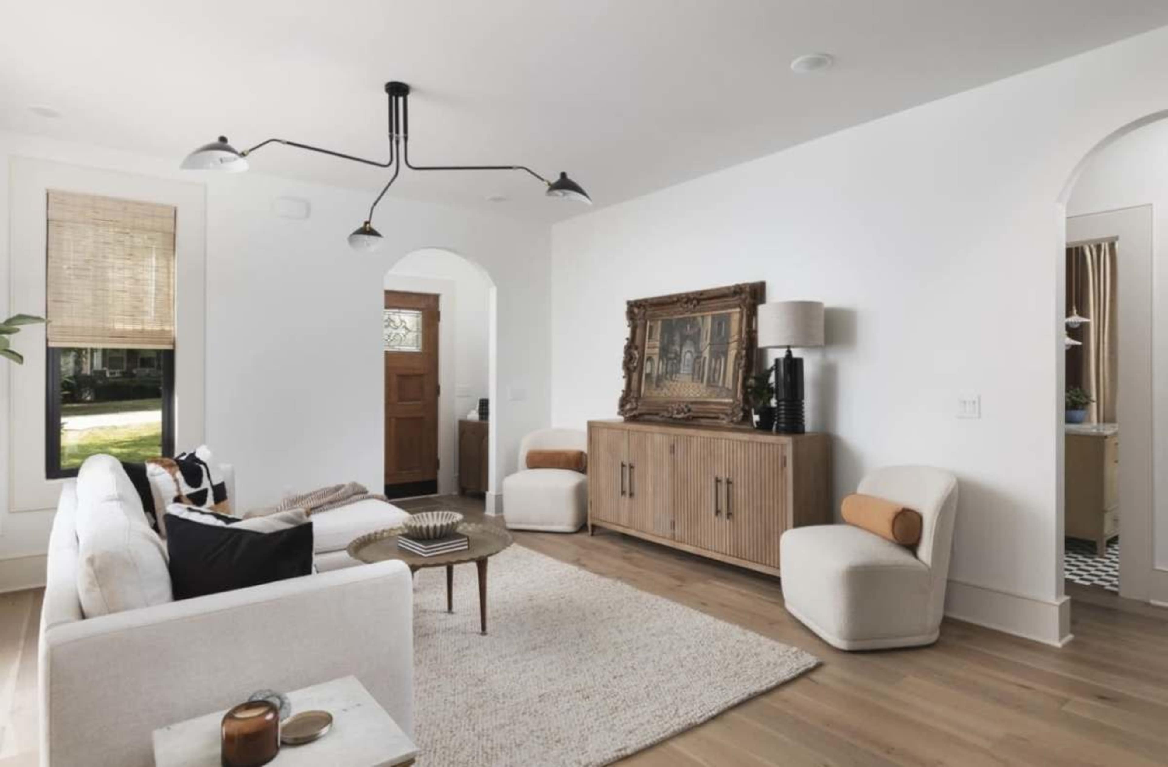 The room features a light-colored sofa, an oval coffee table, and a wooden sideboard, with an art piece displayed on the wall and an entrance visible in the background.