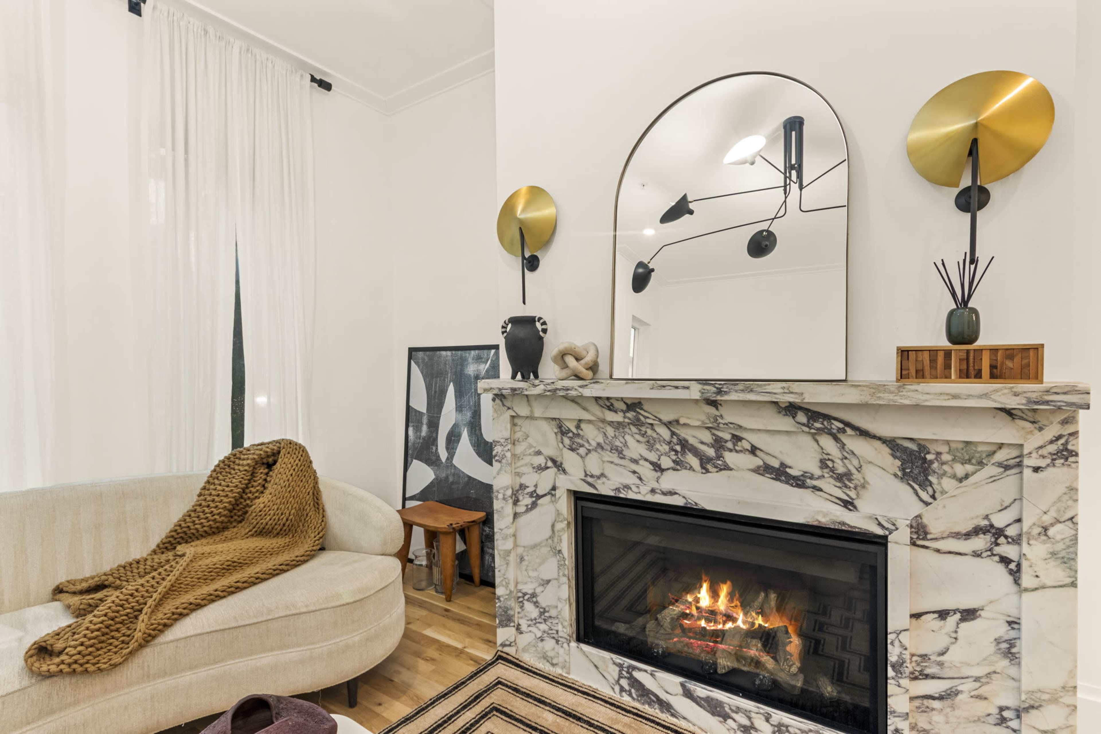 A modern living room features a marble fireplace with a decorative mirror and light fixtures above, accompanied by a cozy sofa draped with a knit blanket.