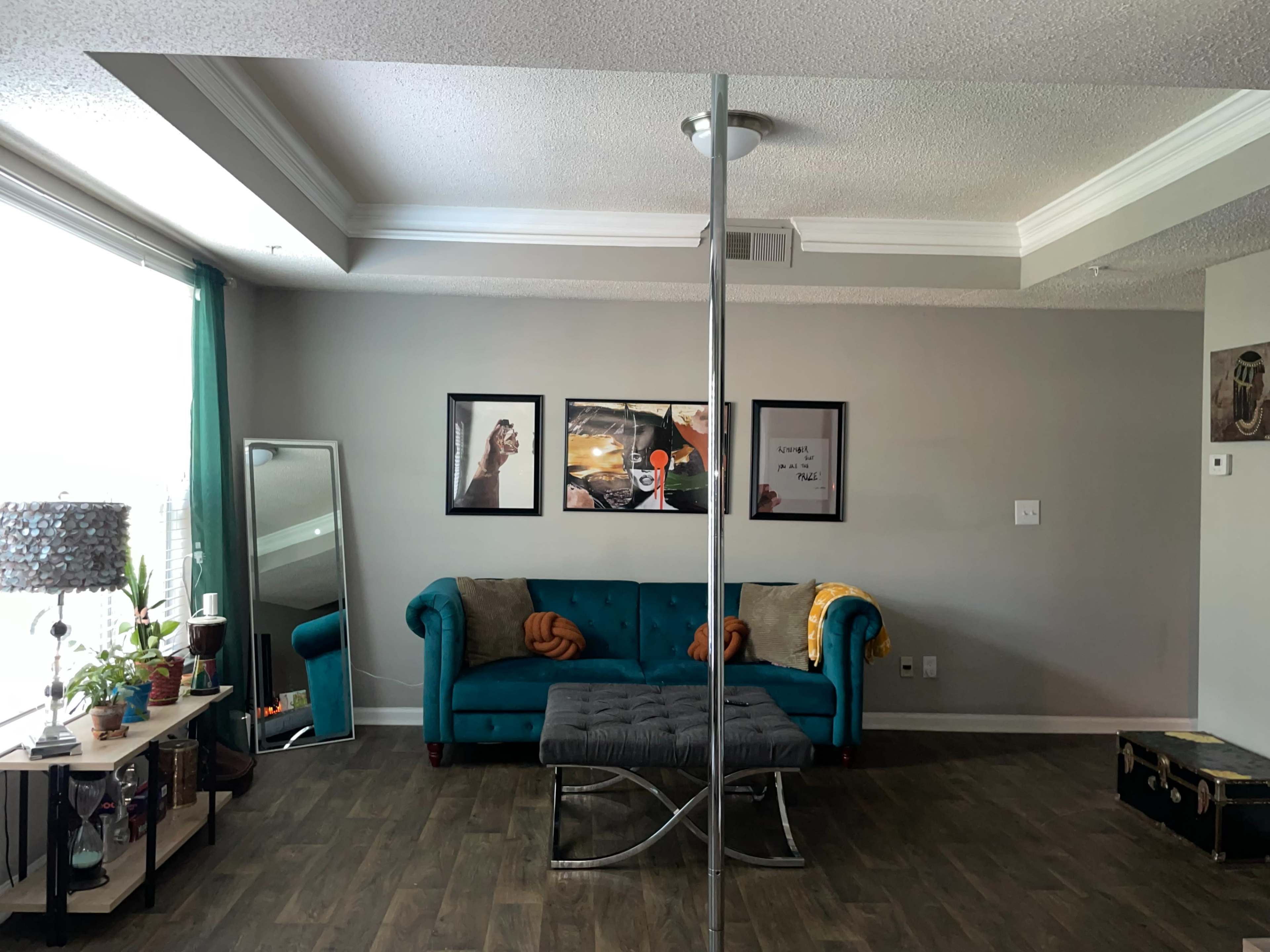 The room features a turquoise velvet couch with decorative pillows, a mirrored accent wall, a coffee table, a lamp, and framed artwork on the gray walls.