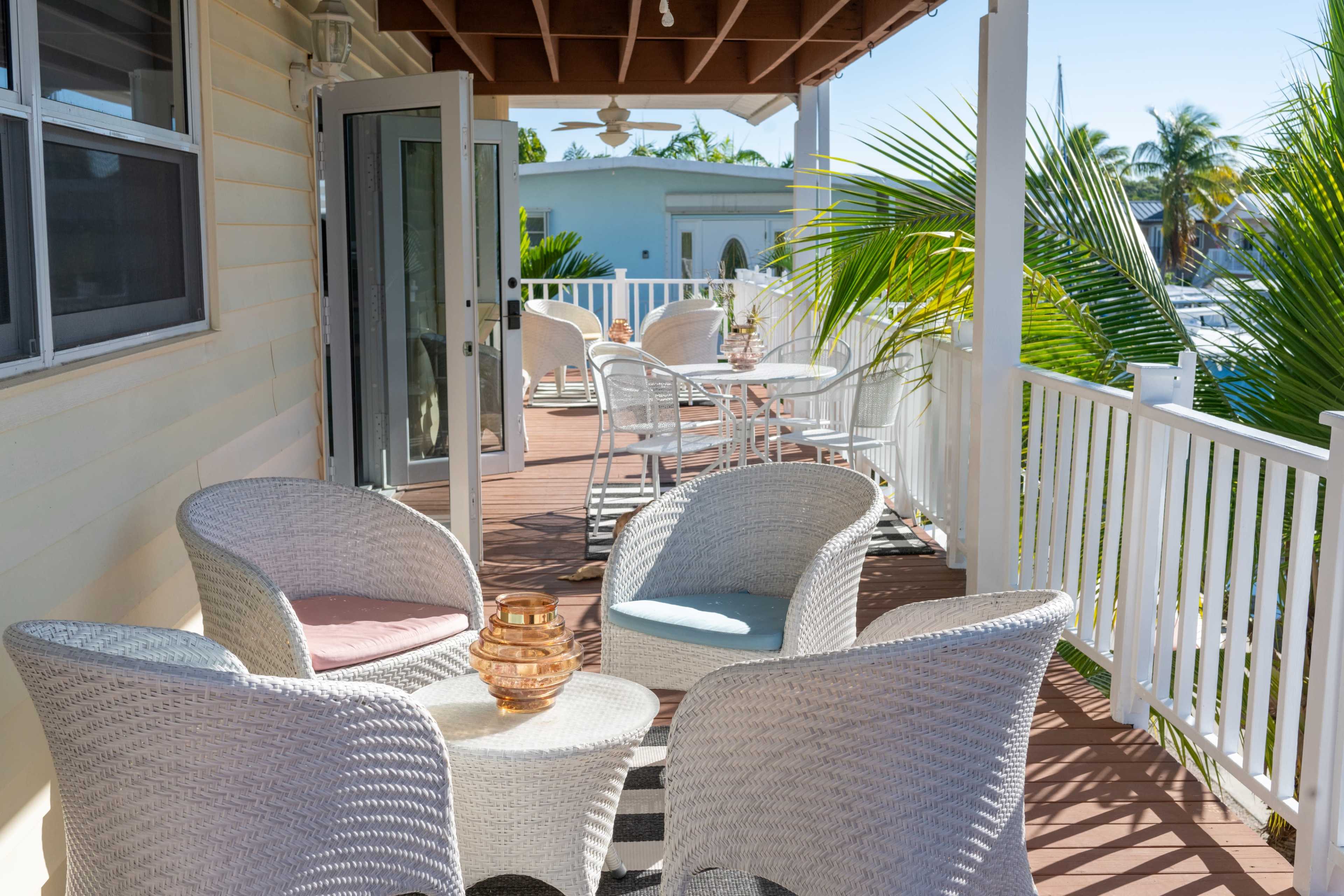 A porch with white wicker chairs and a small table, surrounded by palm trees and sunny outdoor seating areas.