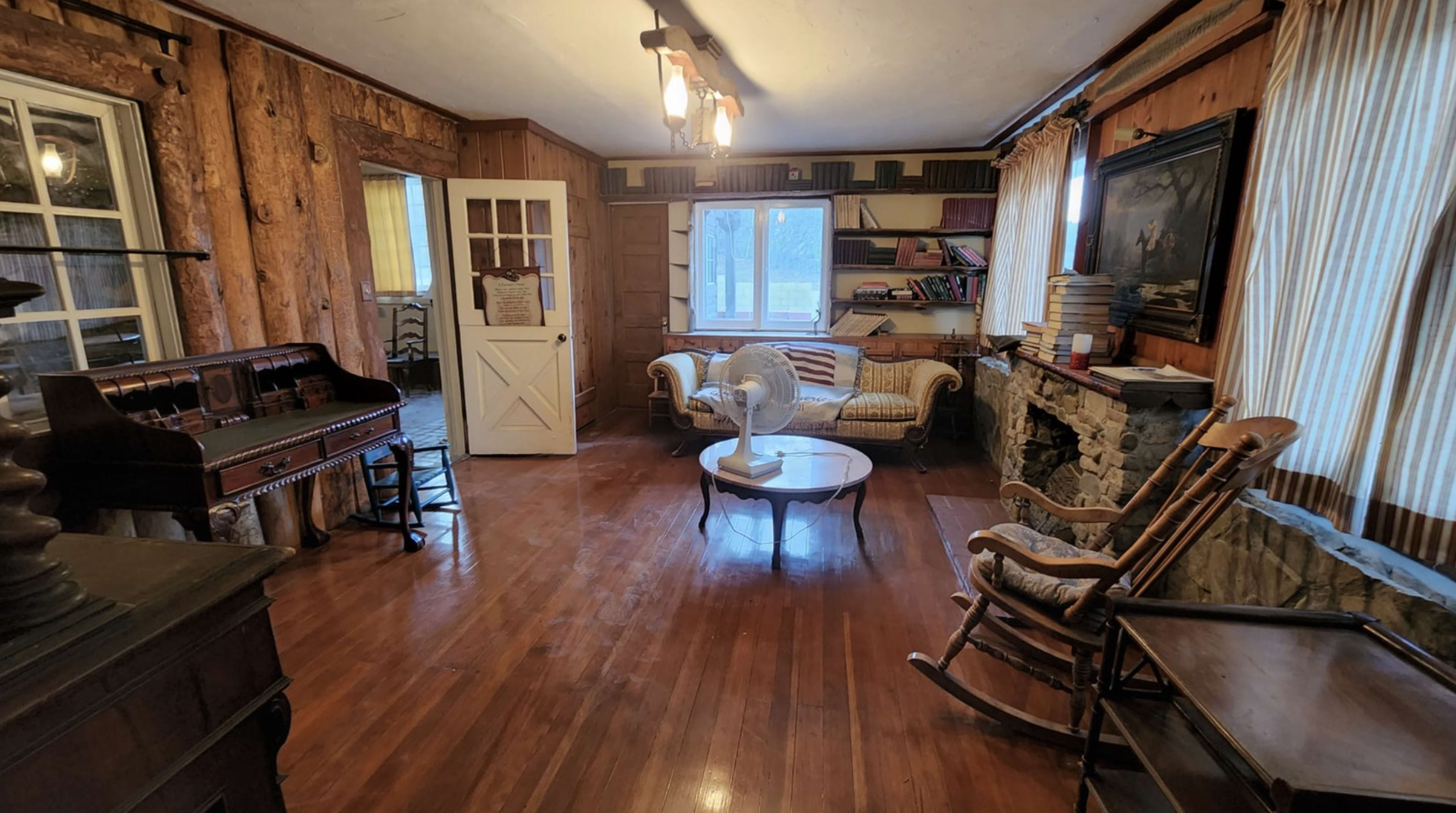 The room features wooden walls, a couch, a rocking chair, a table with a fan, and doorways leading to other spaces.