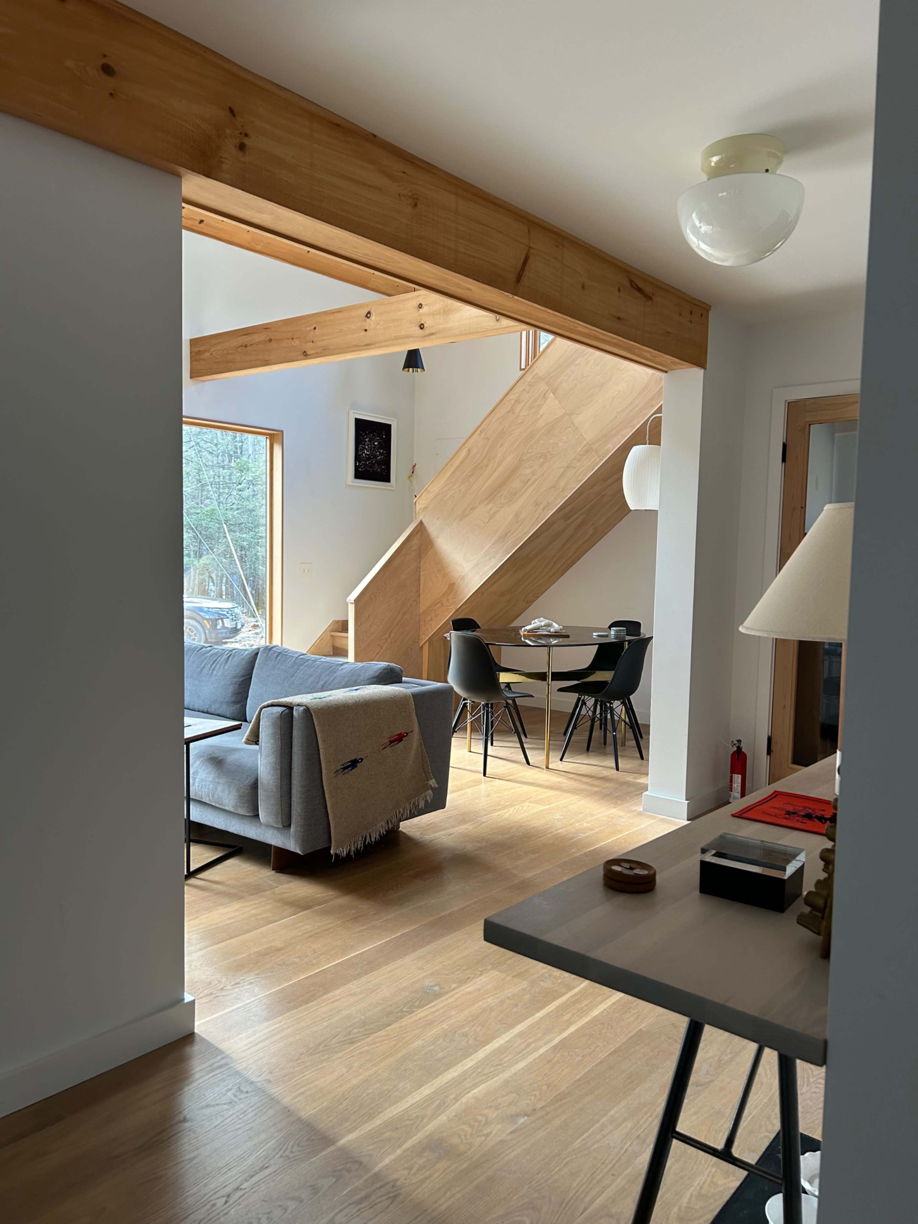 A modern interior space featuring a living area with a gray sofa, a dining area with black chairs, and a staircase with wooden steps.