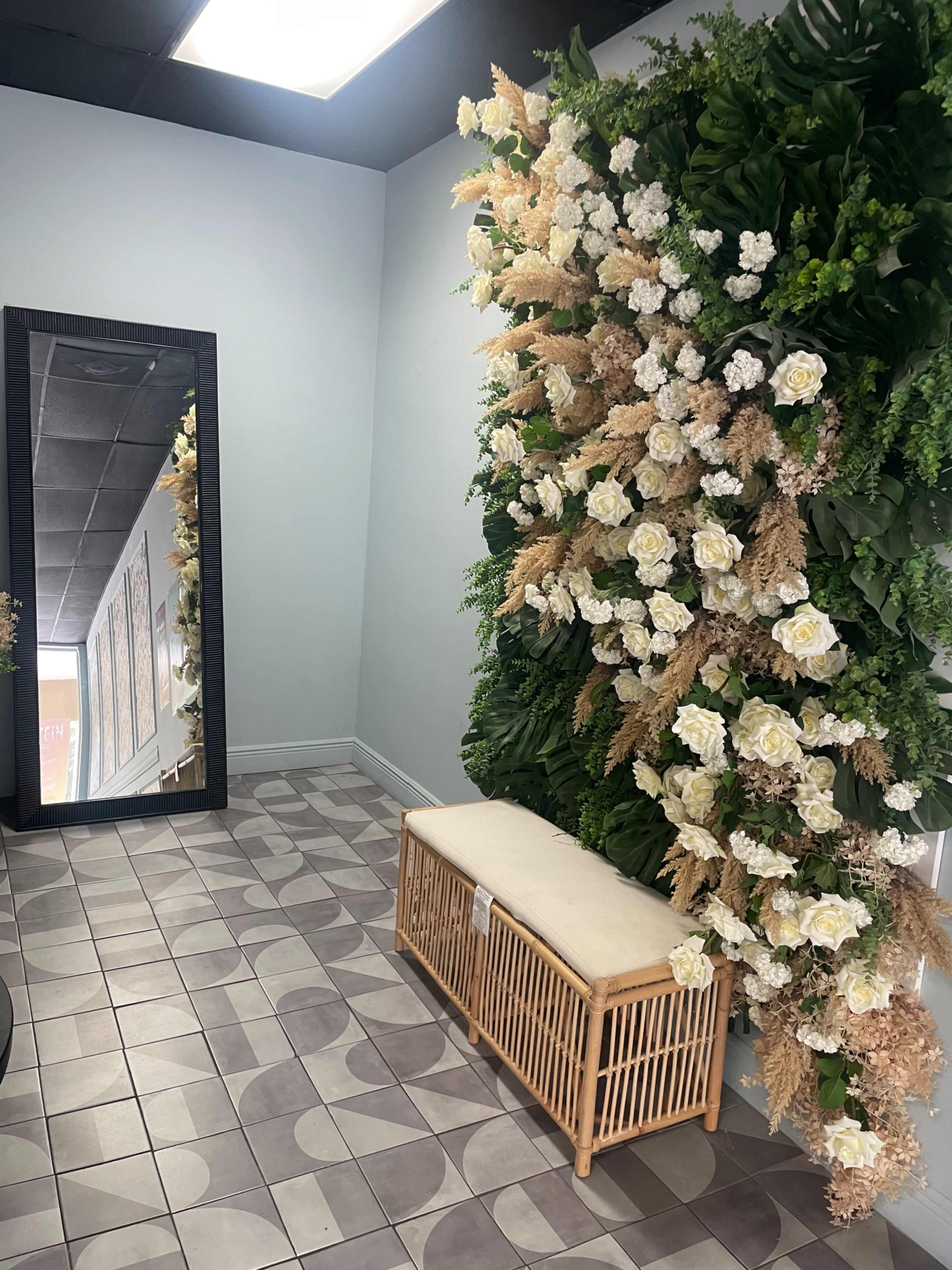 The image shows a hallway with a large mirror on one side and a decorative wall of artificial flowers and greenery on the other, along with a wicker bench on the floor.