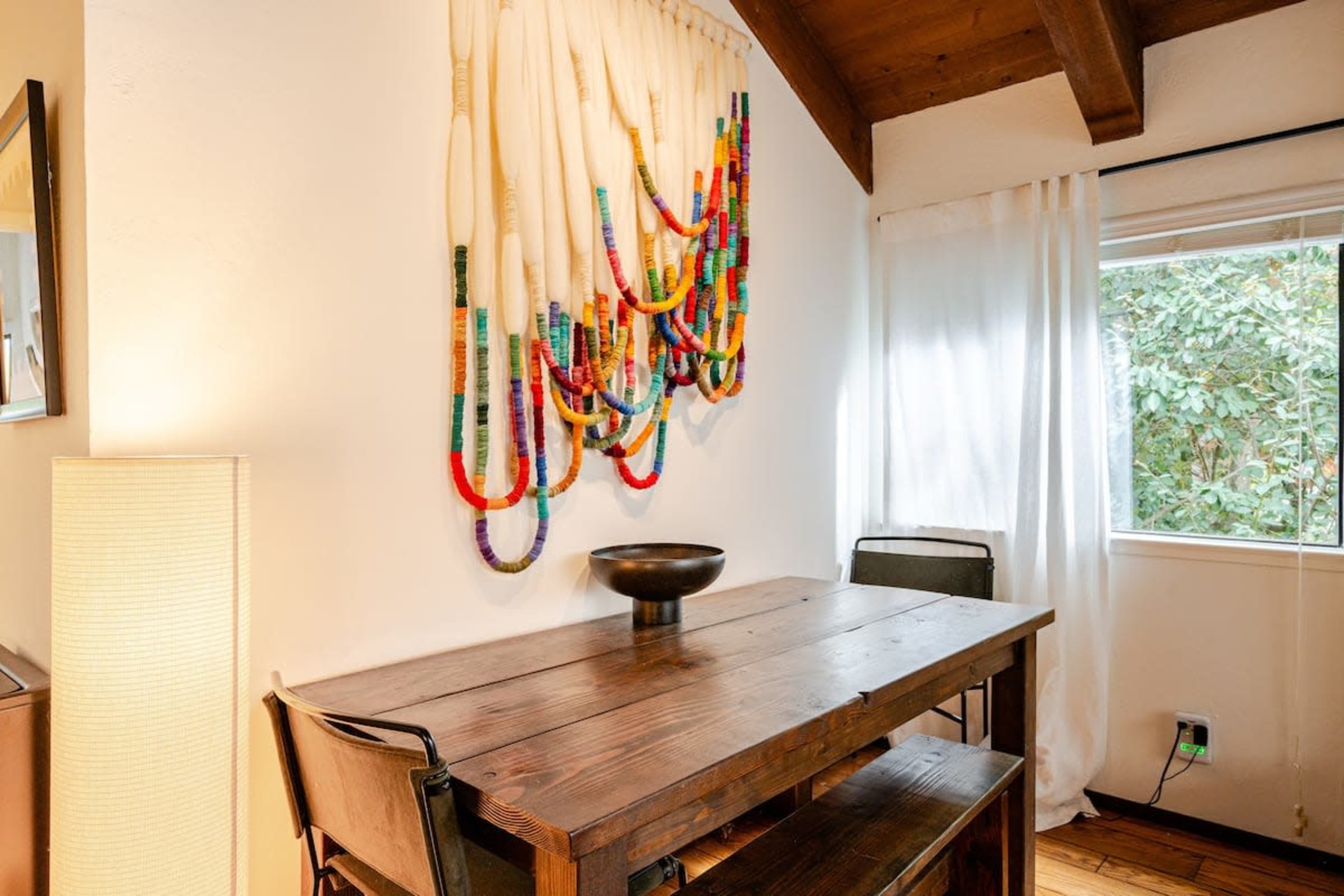 A wooden table with metal chairs is set against a wall adorned with colorful decorative beads, beside a window with sheer curtains.