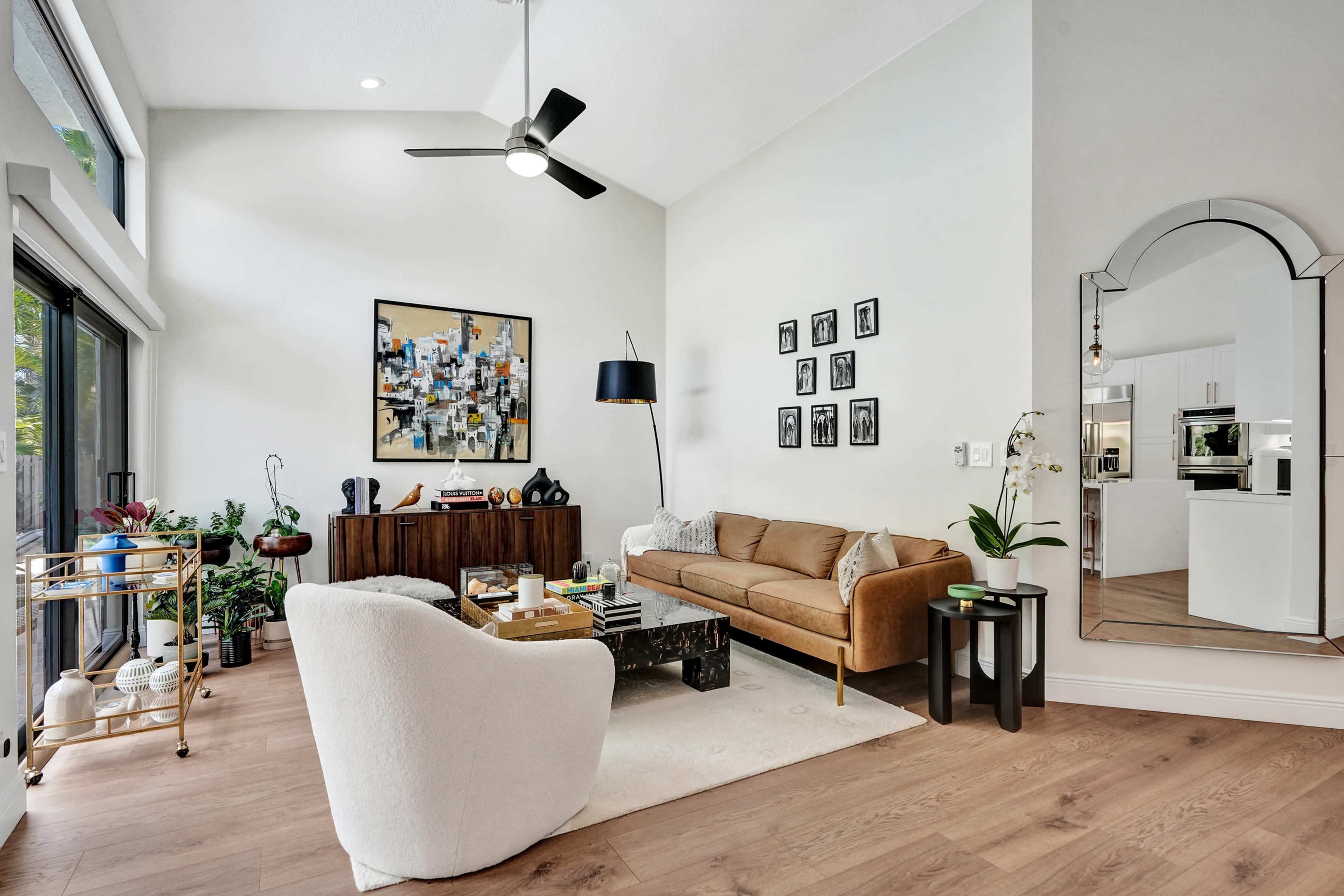 A modern living room features a tan sofa, a white chair, a dark coffee table, and a gallery wall with black-and-white photos, surrounded by greenery.