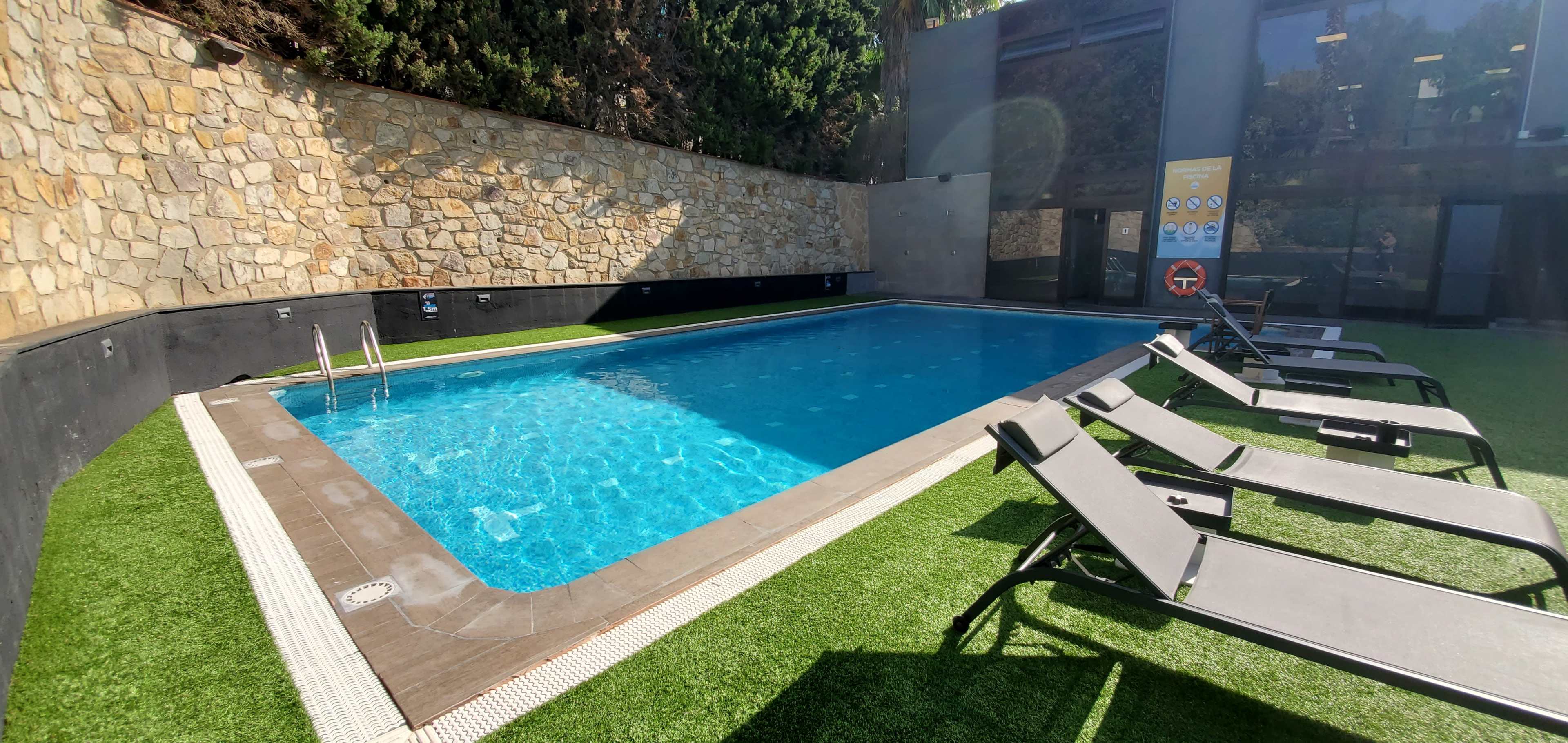 A rectangular swimming pool is surrounded by lounge chairs and a stone wall in a sunlit area.
