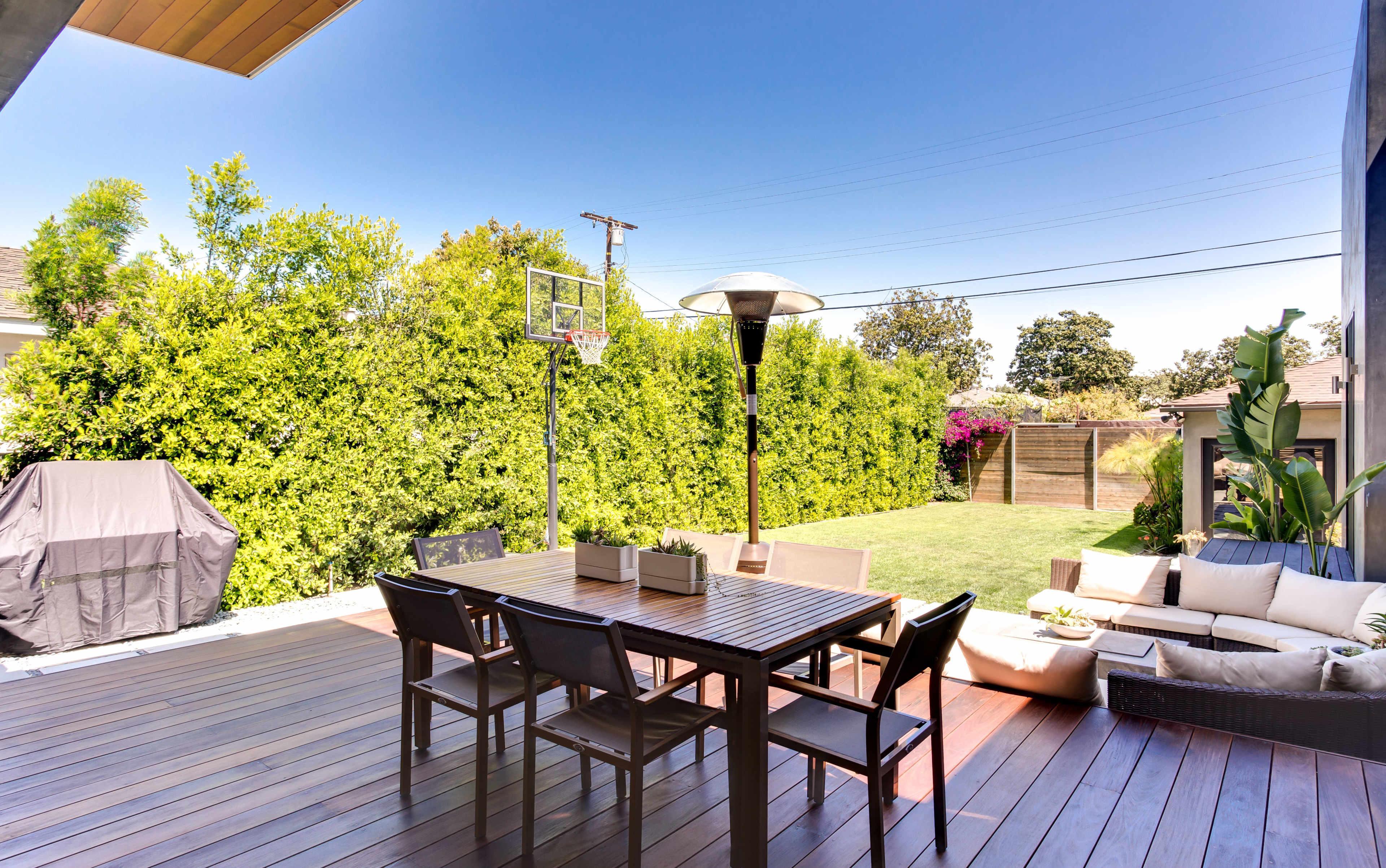 A backyard patio with a wooden dining table and chairs, a basketball hoop, a barbecue grill, and a lawn bordered by dense greenery.