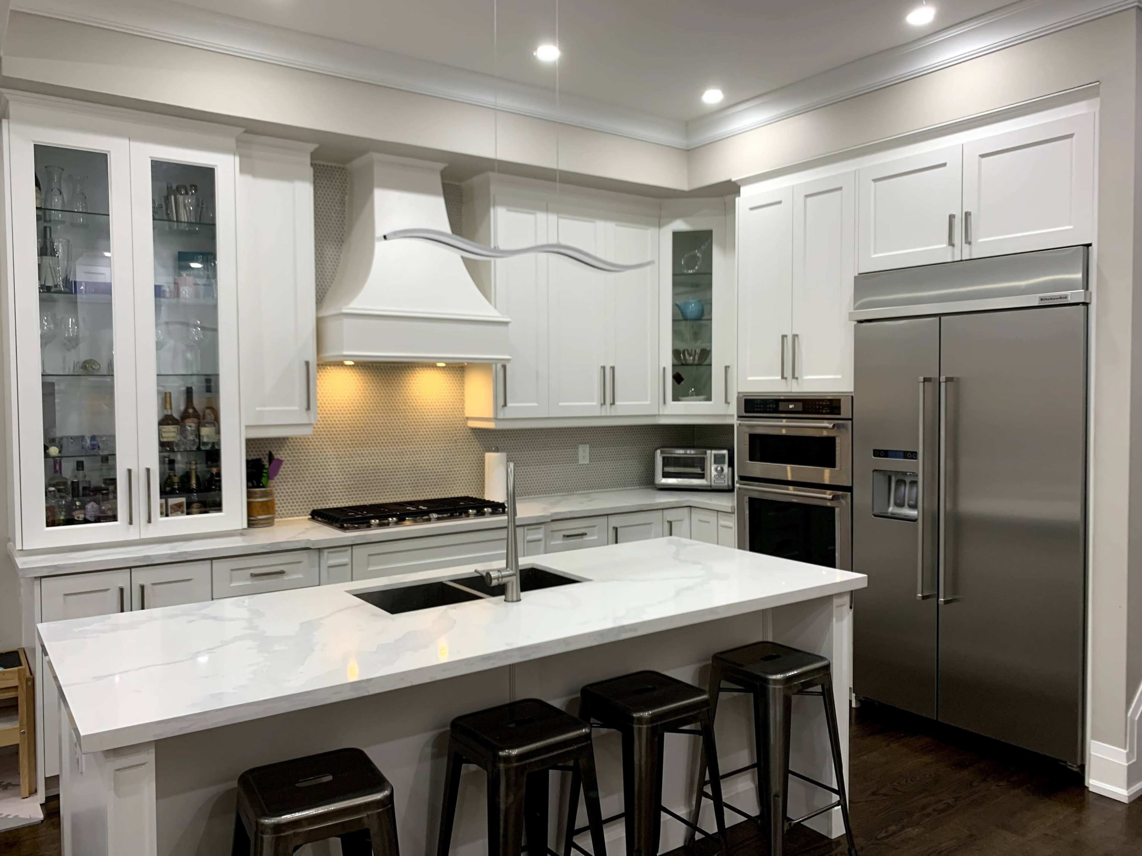 A modern kitchen with white cabinets, a large island with bar stools, stainless steel appliances, and a hood above the stove.