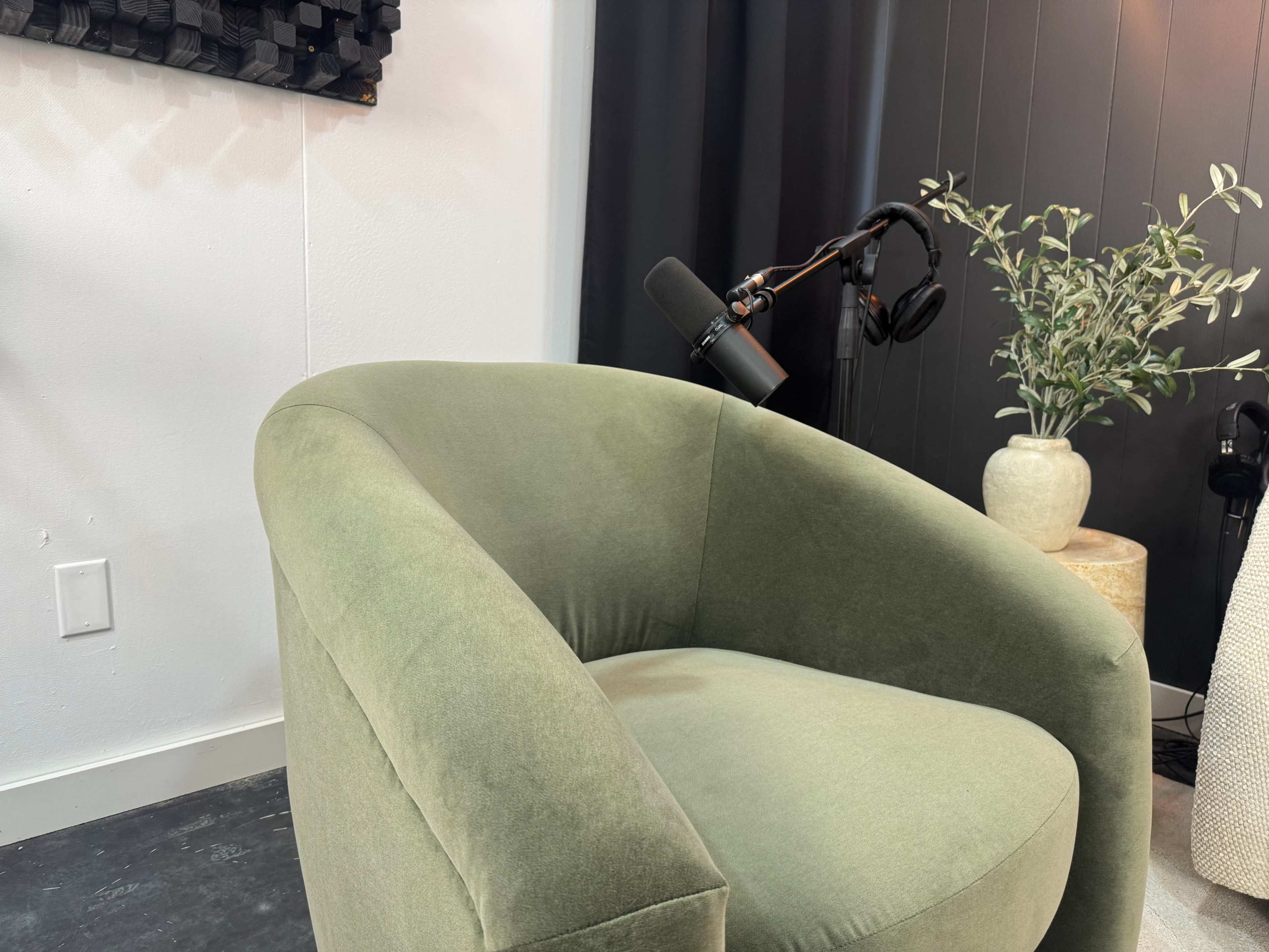 The image shows a green upholstered chair beside a table with a plant and a microphone mounted on an arm in a minimalist room setting.