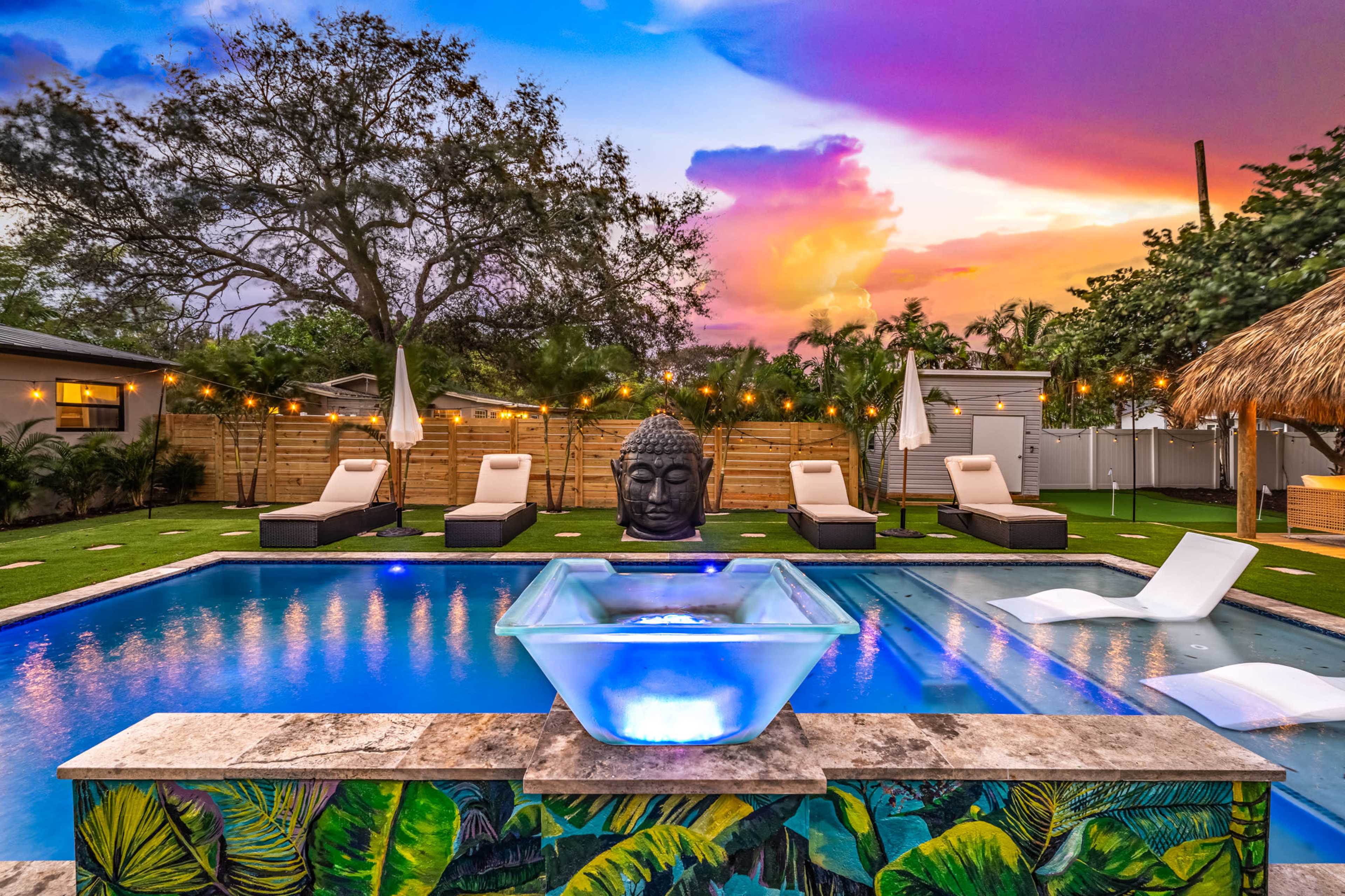 A modern backyard features a swimming pool with a fountain, lounge chairs, a large Buddha statue, and vibrant sunset skies above.