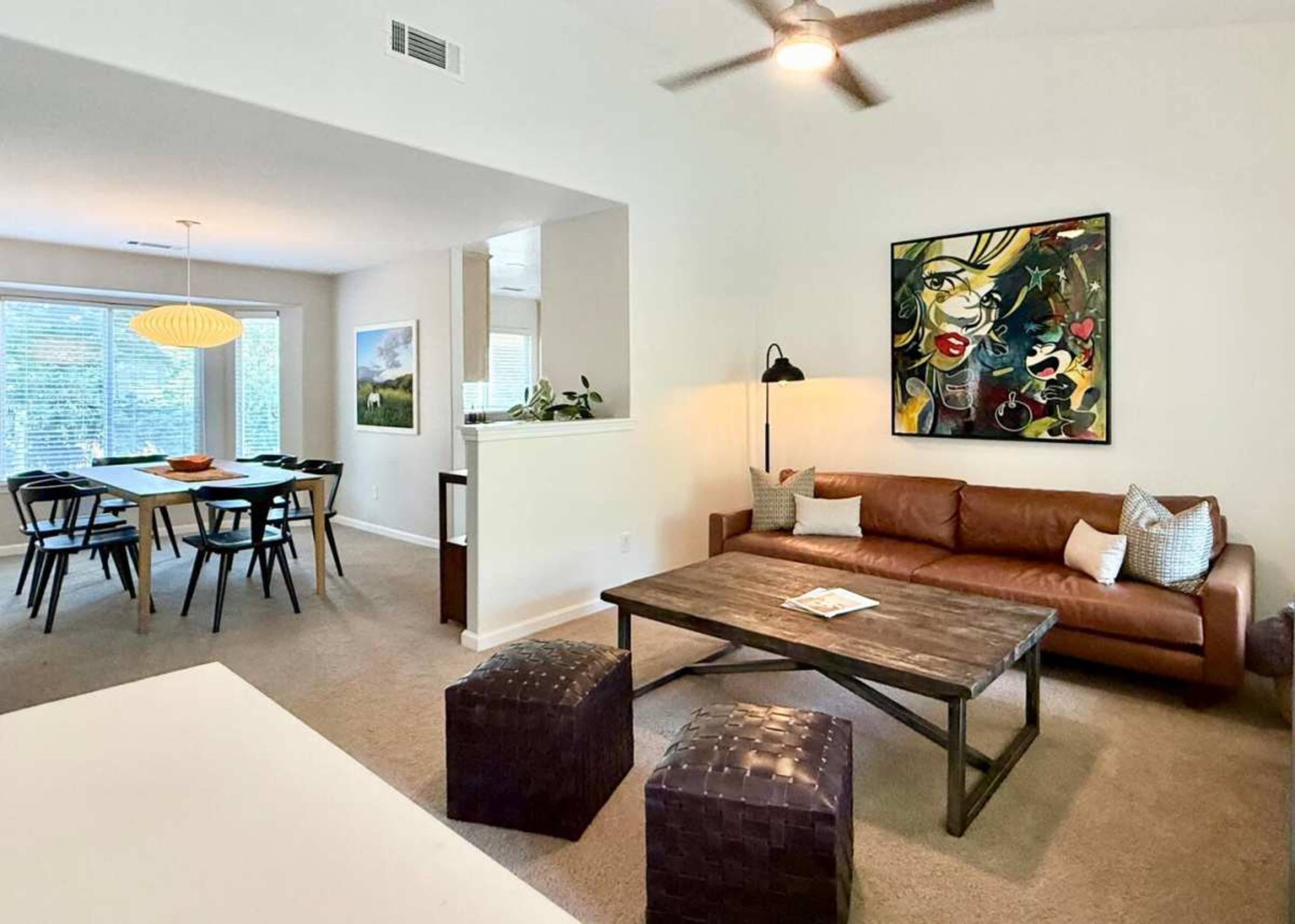 The image shows a modern living room with a brown leather sofa, a wooden coffee table, and a dining area in the background featuring a long table and chairs.