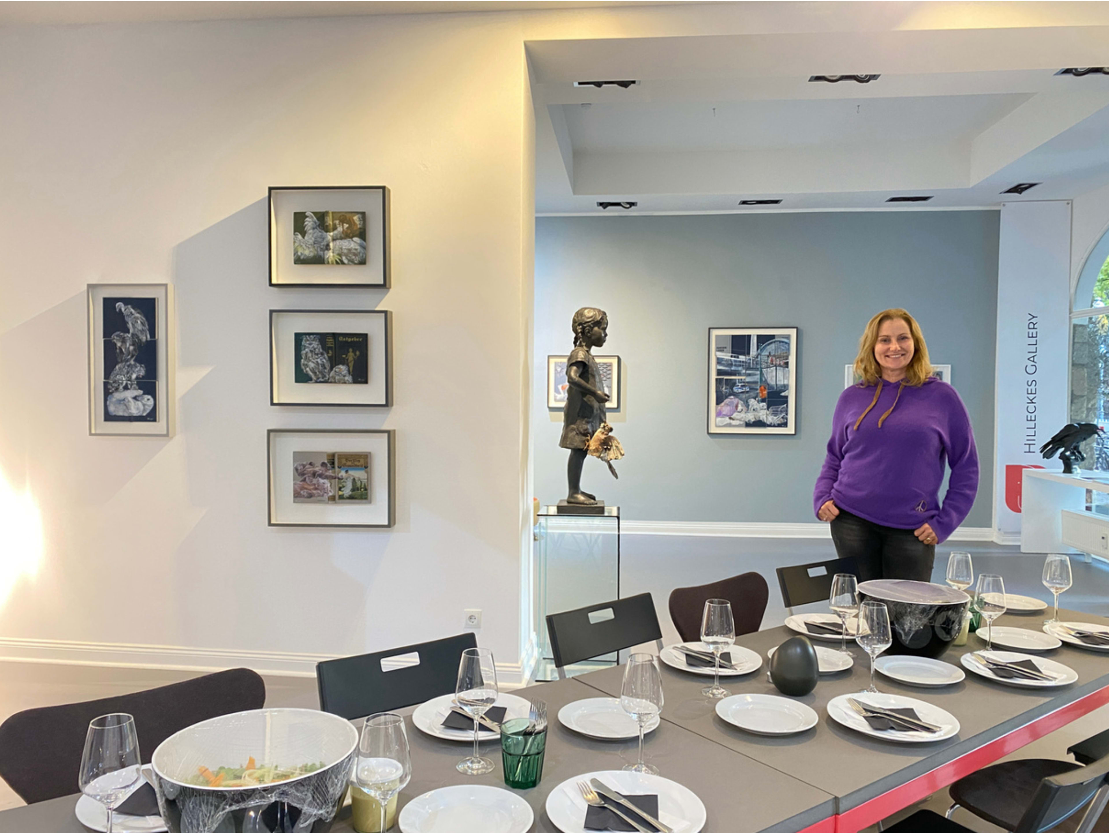 A woman in a purple sweatshirt stands beside a well-set dining table in an art gallery featuring framed artwork on the walls.