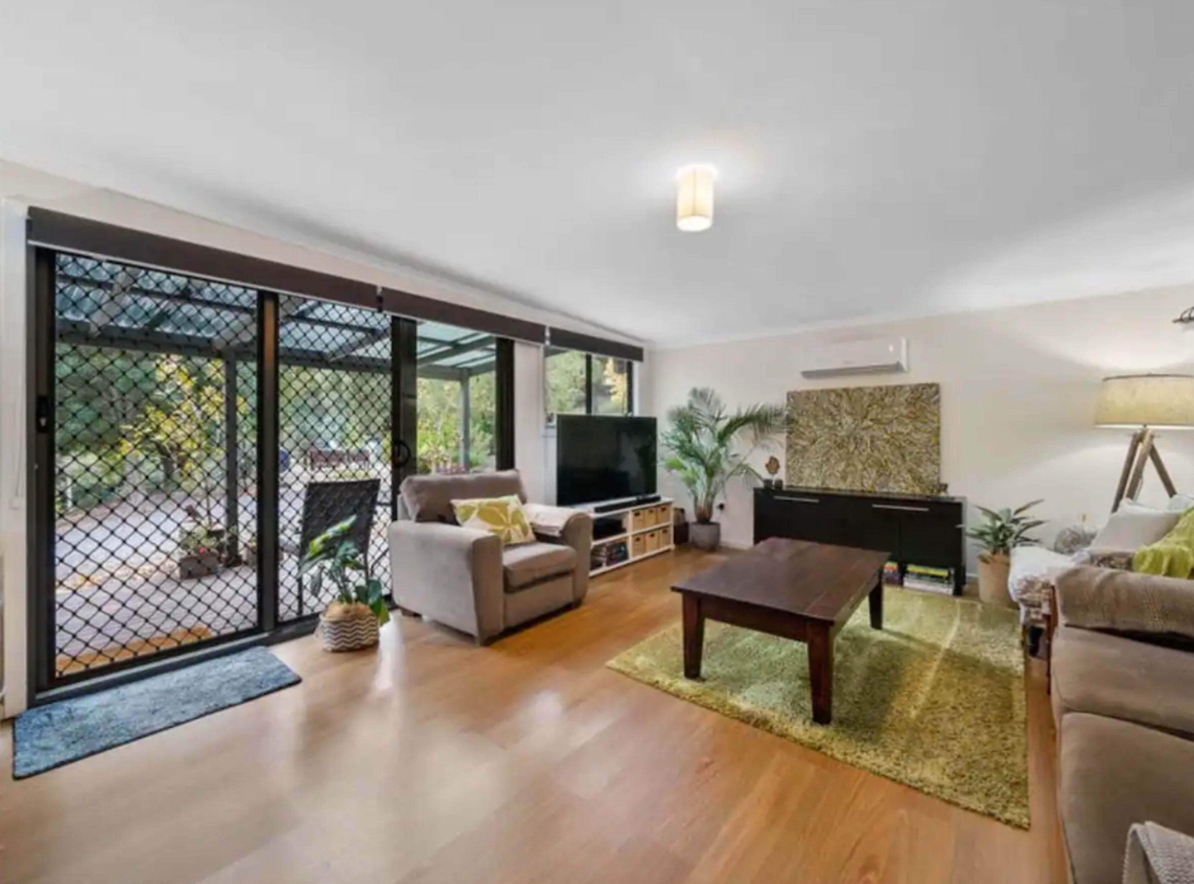 The living room features a wooden table surrounded by sofas, with large glass doors leading to an outdoor area.