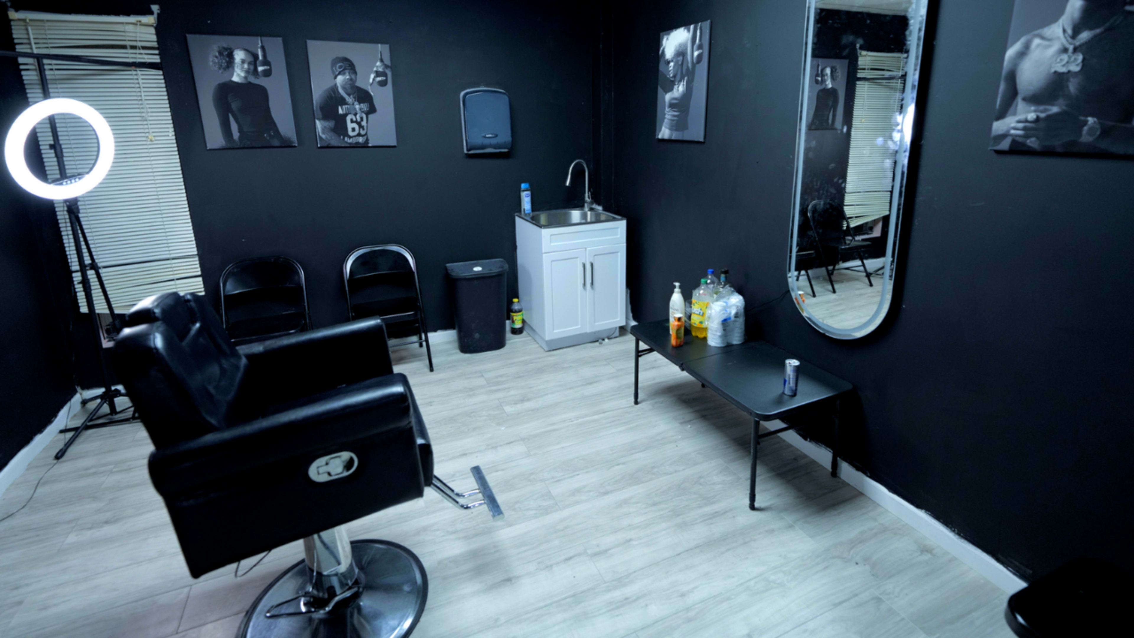 The image shows a black-walled barbershop with black chairs, a sink, a mirror, and a table with bottles and a can.