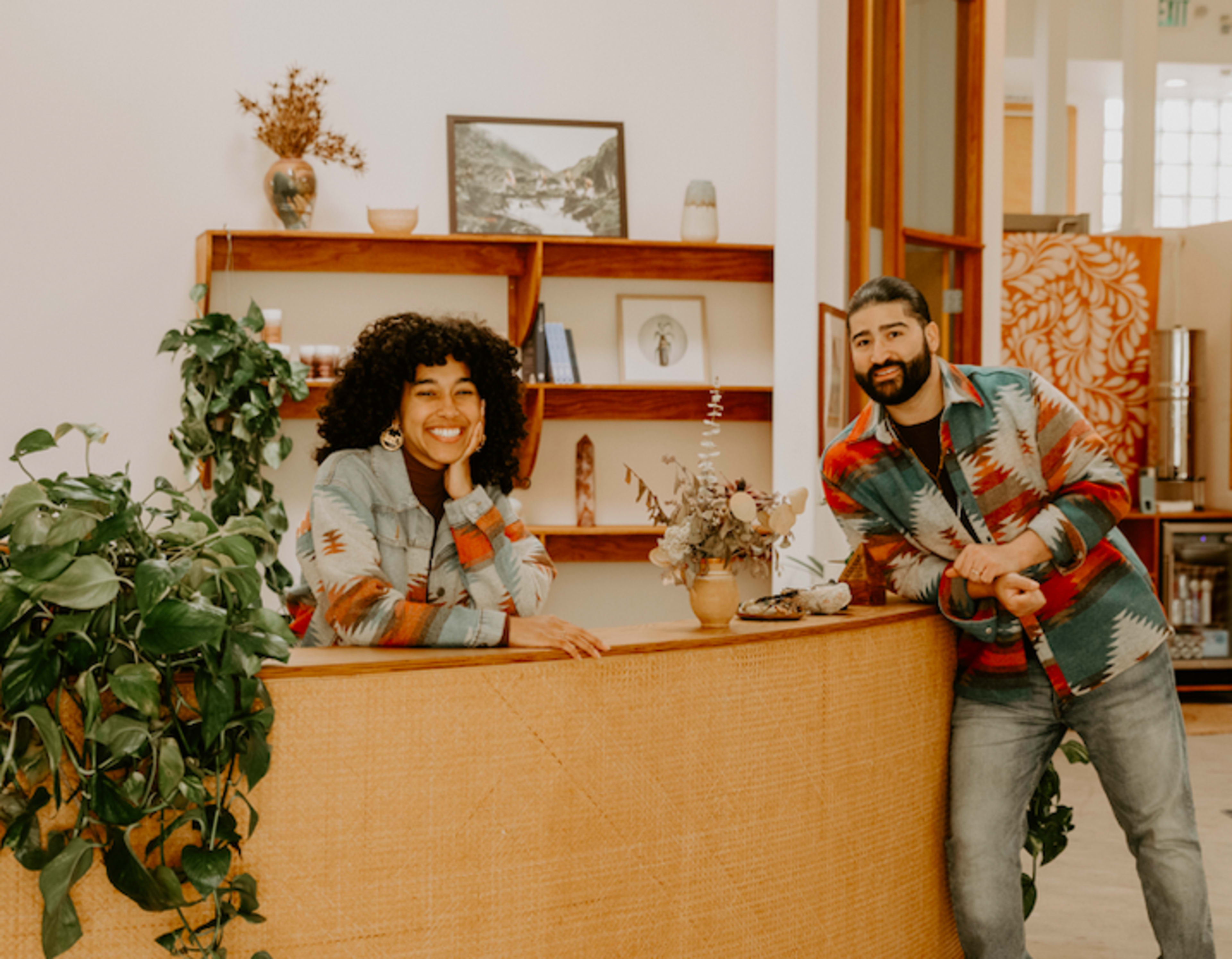 A man and a woman stand at a reception desk in a bright, modern space decorated with plants and shelves of decorative items.