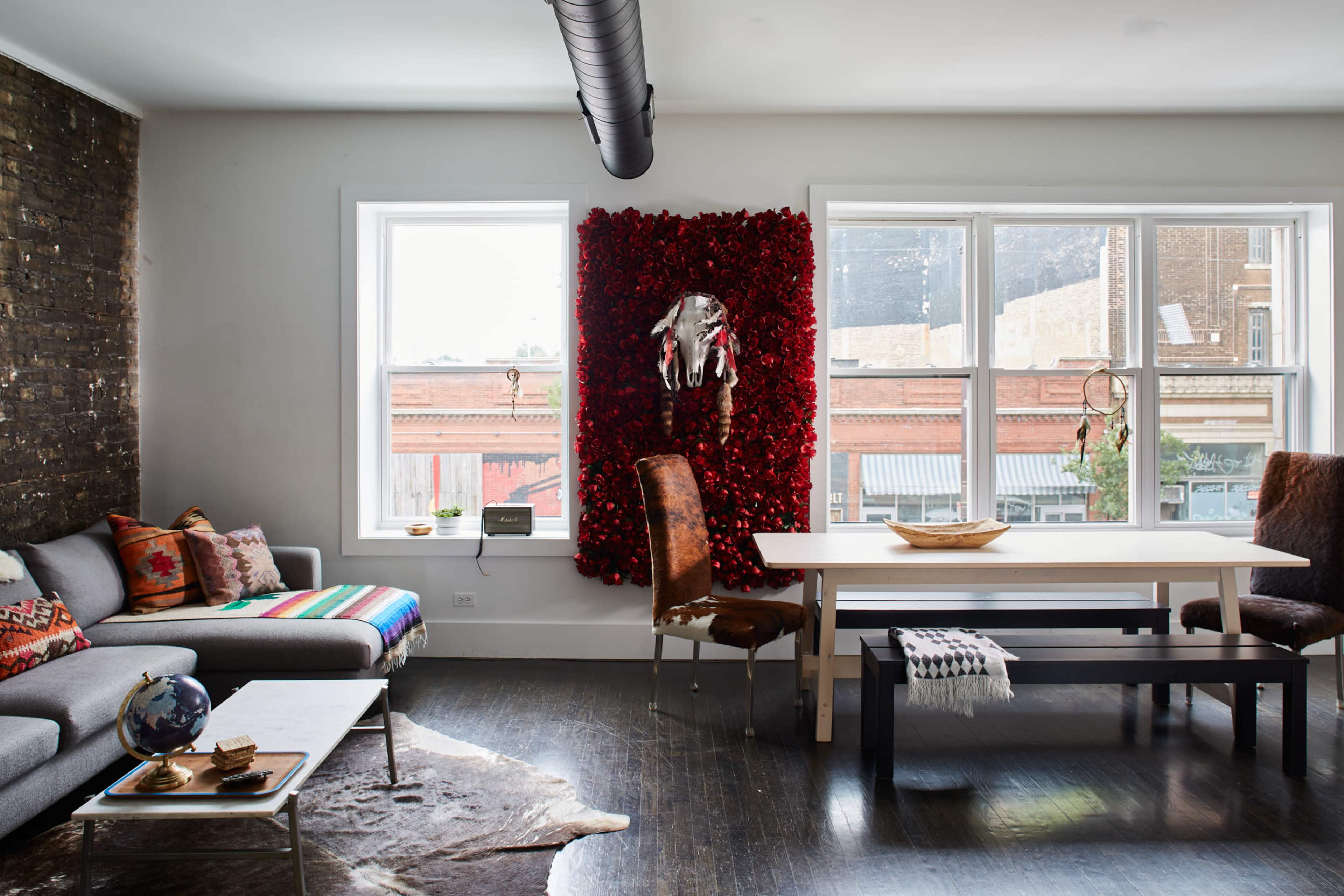 A modern living room features a sectional sofa, a large white dining table, and a wall adorned with red floral decor and an animal skull, complemented by large windows overlooking a brick street.