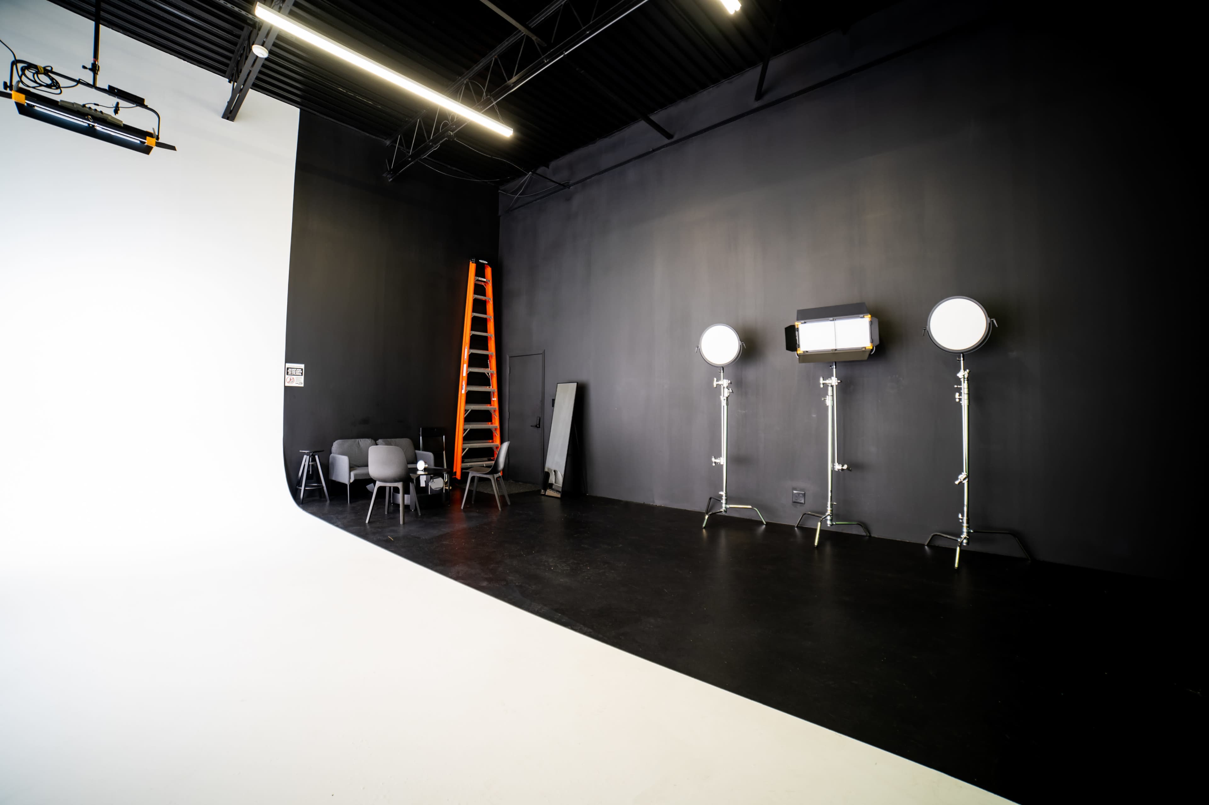 A photography studio features a white backdrop on one side, black walls, a ladder, lighting equipment, and a few chairs.