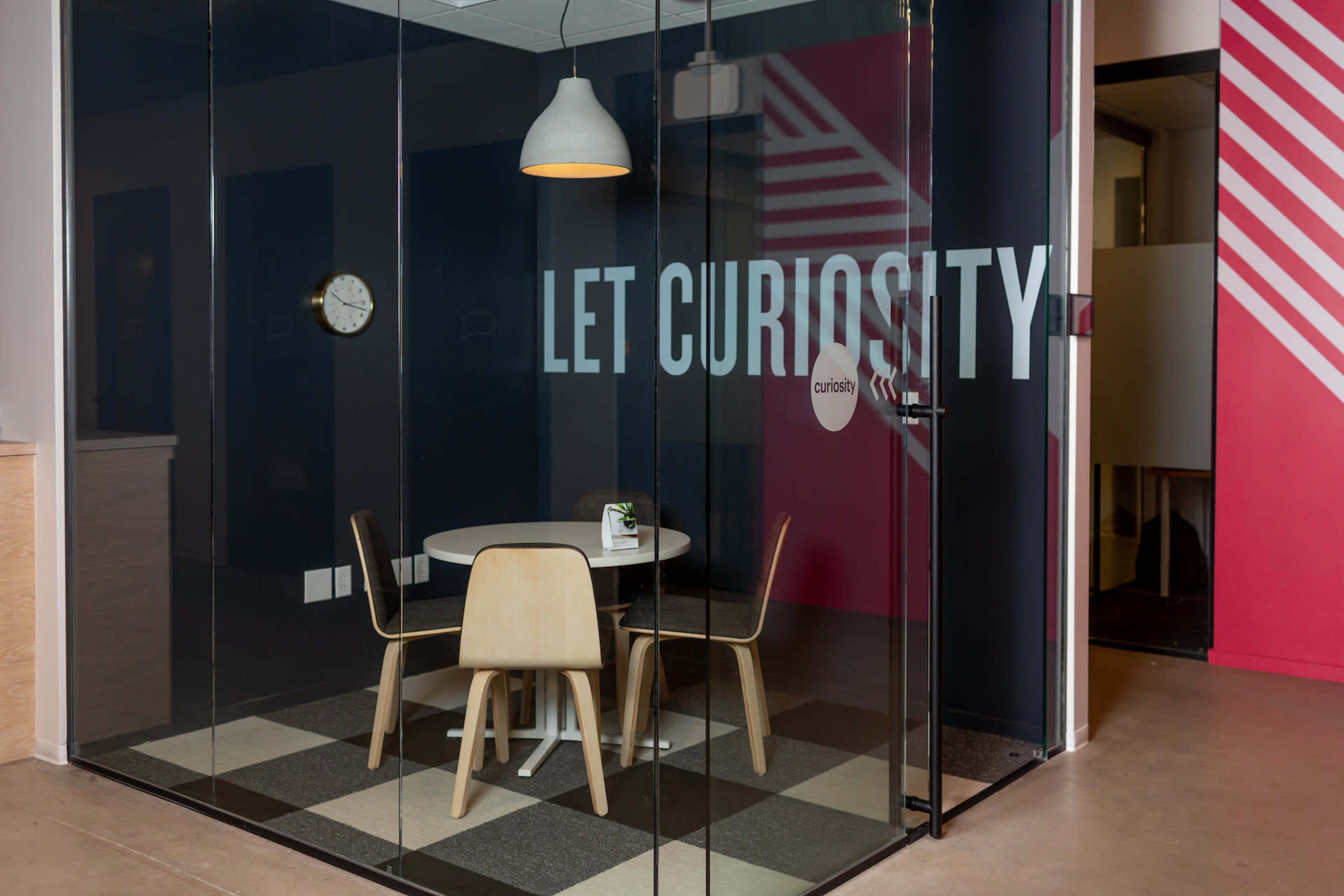 A modern glass conference room with a round table and wooden chairs, featuring a wall with the phrase "LET CURIOSITY" prominently displayed.