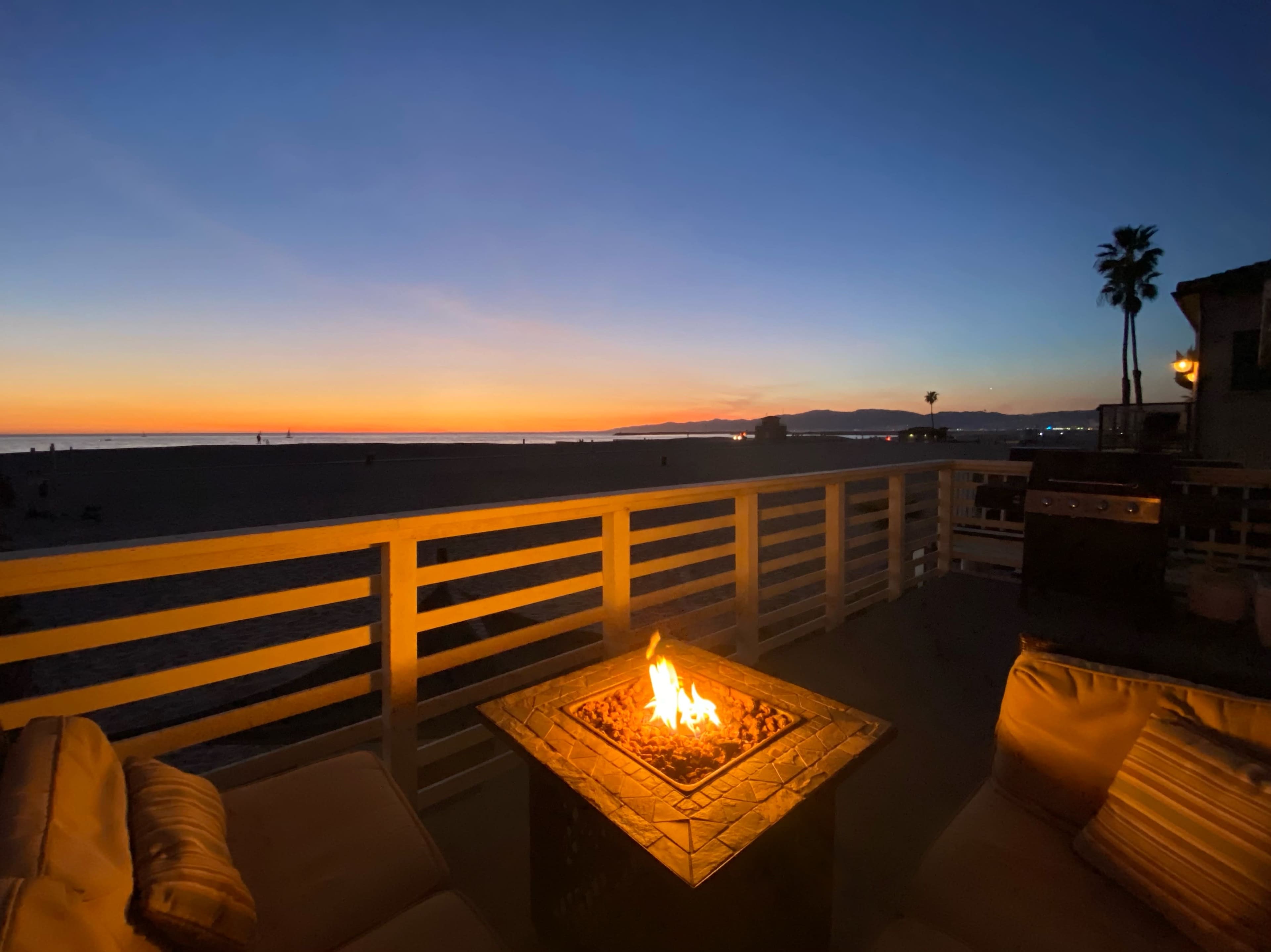 A fire pit glows on a patio overlooking a beach at sunset, with silhouettes of palm trees and distant hills.