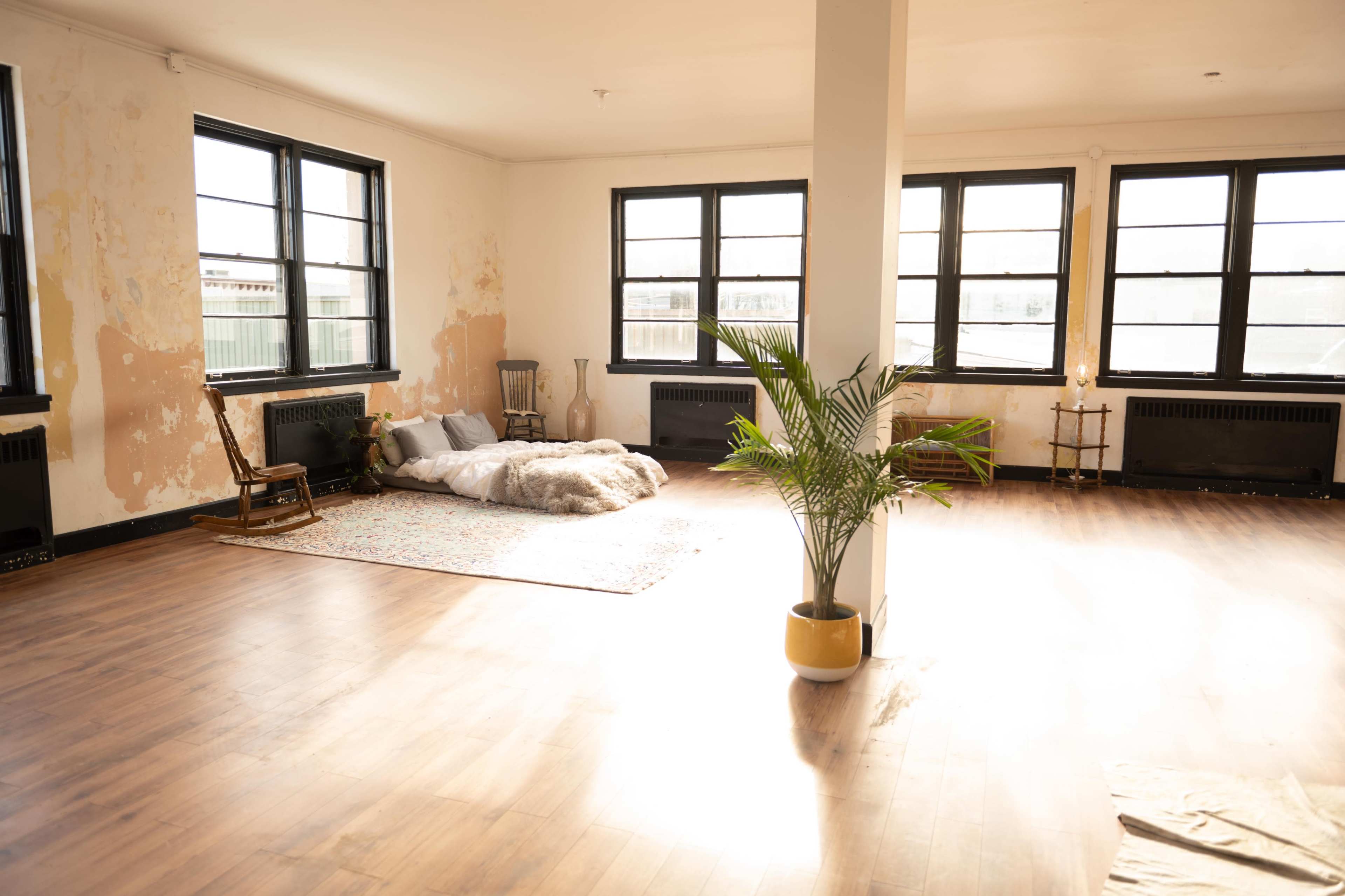 A spacious, partially furnished room with wooden floors, large windows, a soft area for sitting, and a potted plant.