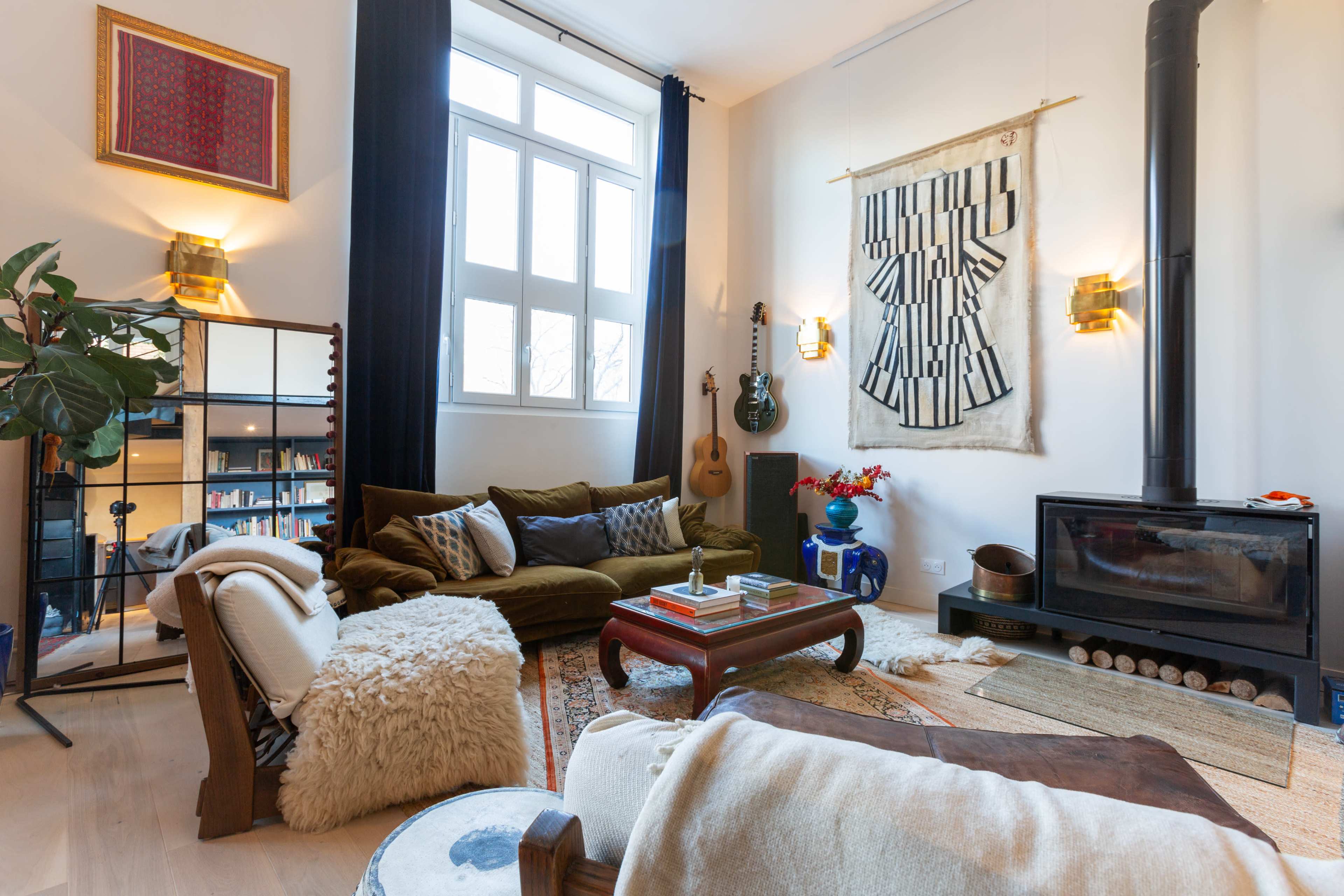 A cozy living room featuring a green sofa, a guitar on the wall, and a wood-burning stove, along with decorative art and plants.