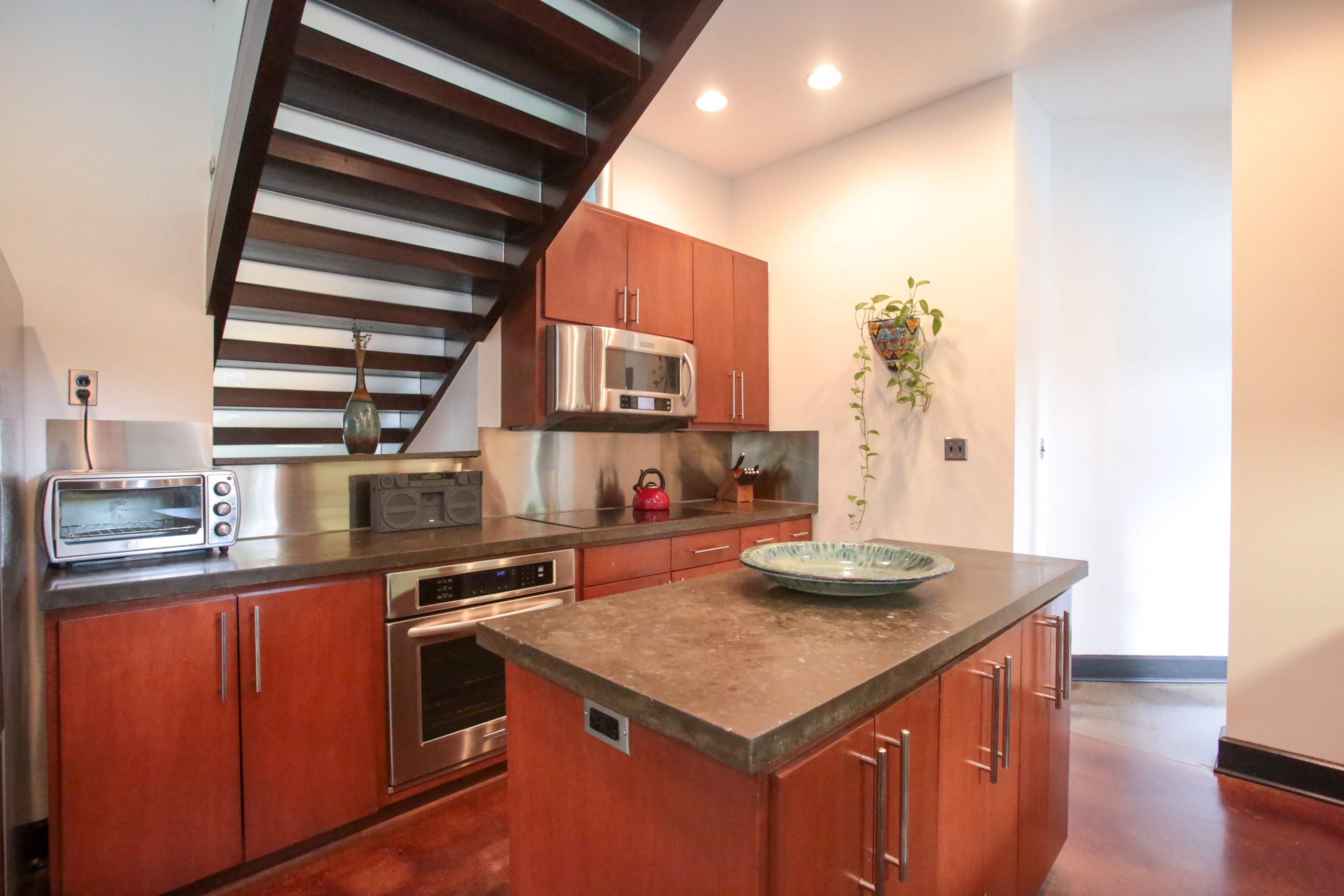 A modern kitchen features dark wood cabinets, stainless steel appliances, and a concrete countertop, with a staircase leading to an upper level.