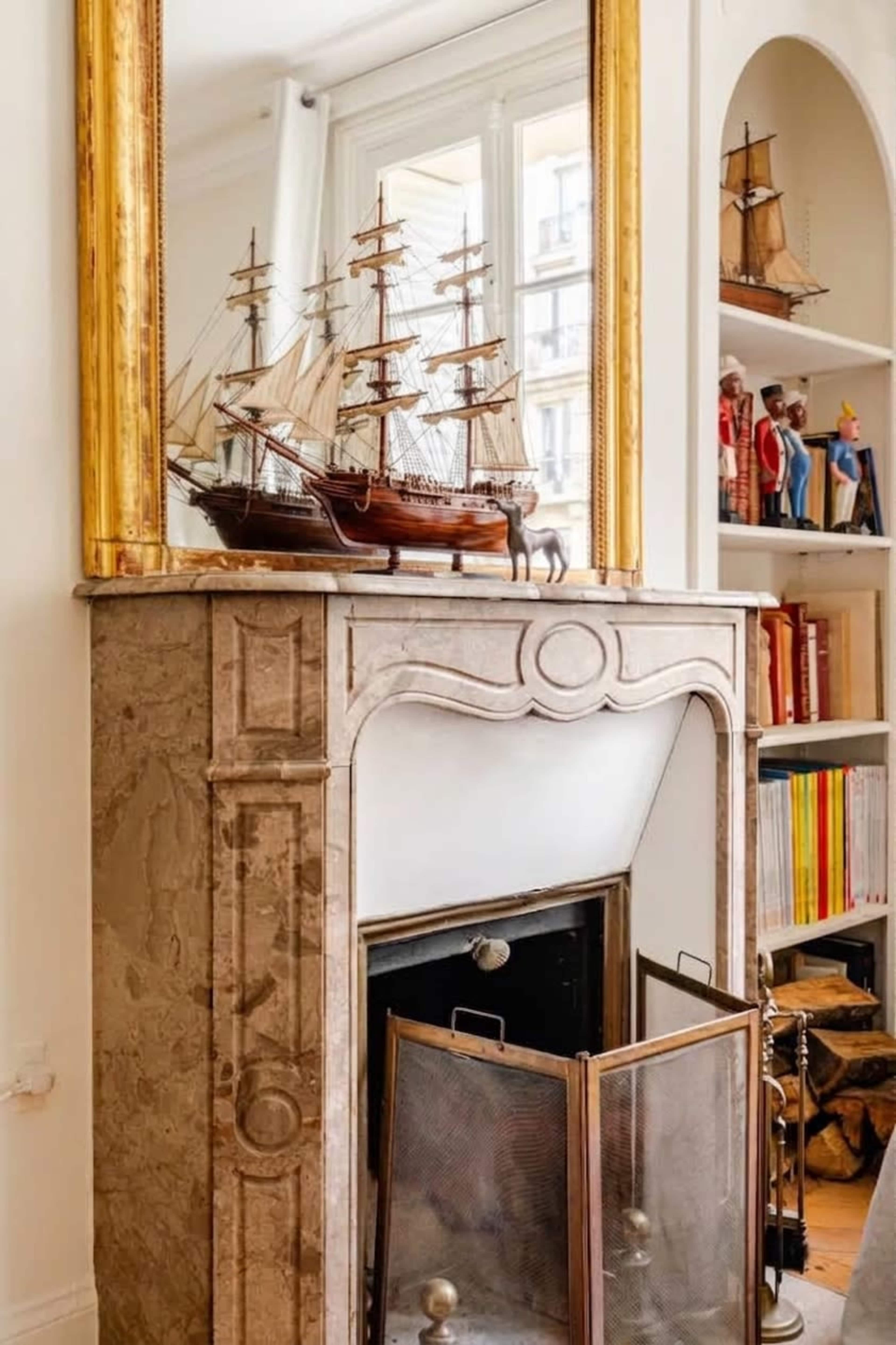 The image features a decorative fireplace with a marble surround, a large mirror above it, and model ships displayed on top.