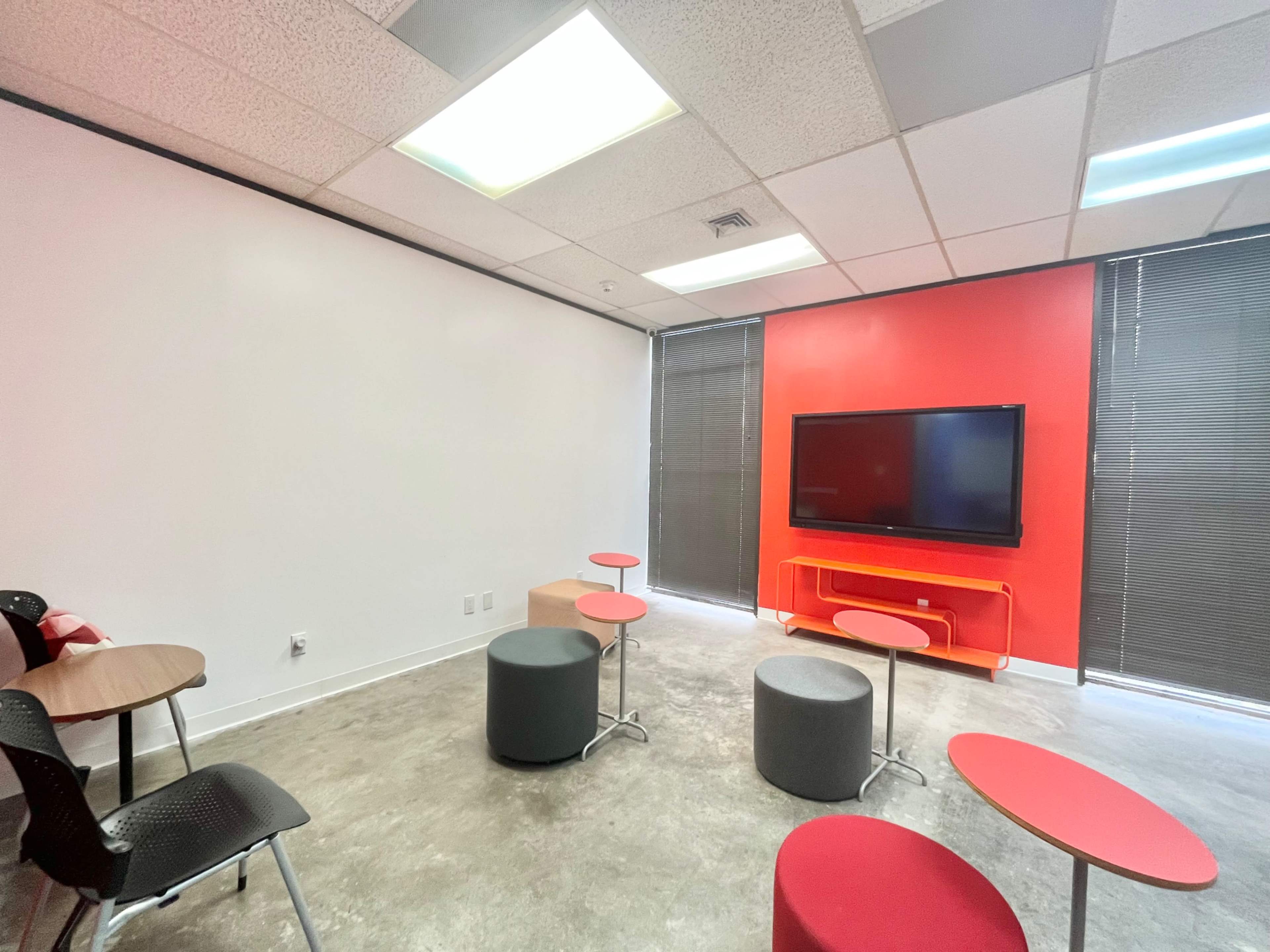 The image shows a small meeting room with a red accent wall, a flat-screen TV mounted on the wall, and circular tables and stools in various shades of gray and red.